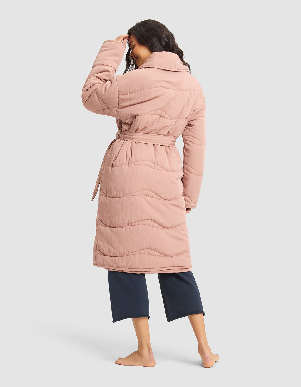 A woman stands barefoot, facing away, wearing the Cozy Earth Women's Quilted House Coat in light pink with a belt and wide collar over dark cropped pants, set against a plain light background. |Color:Rosewood