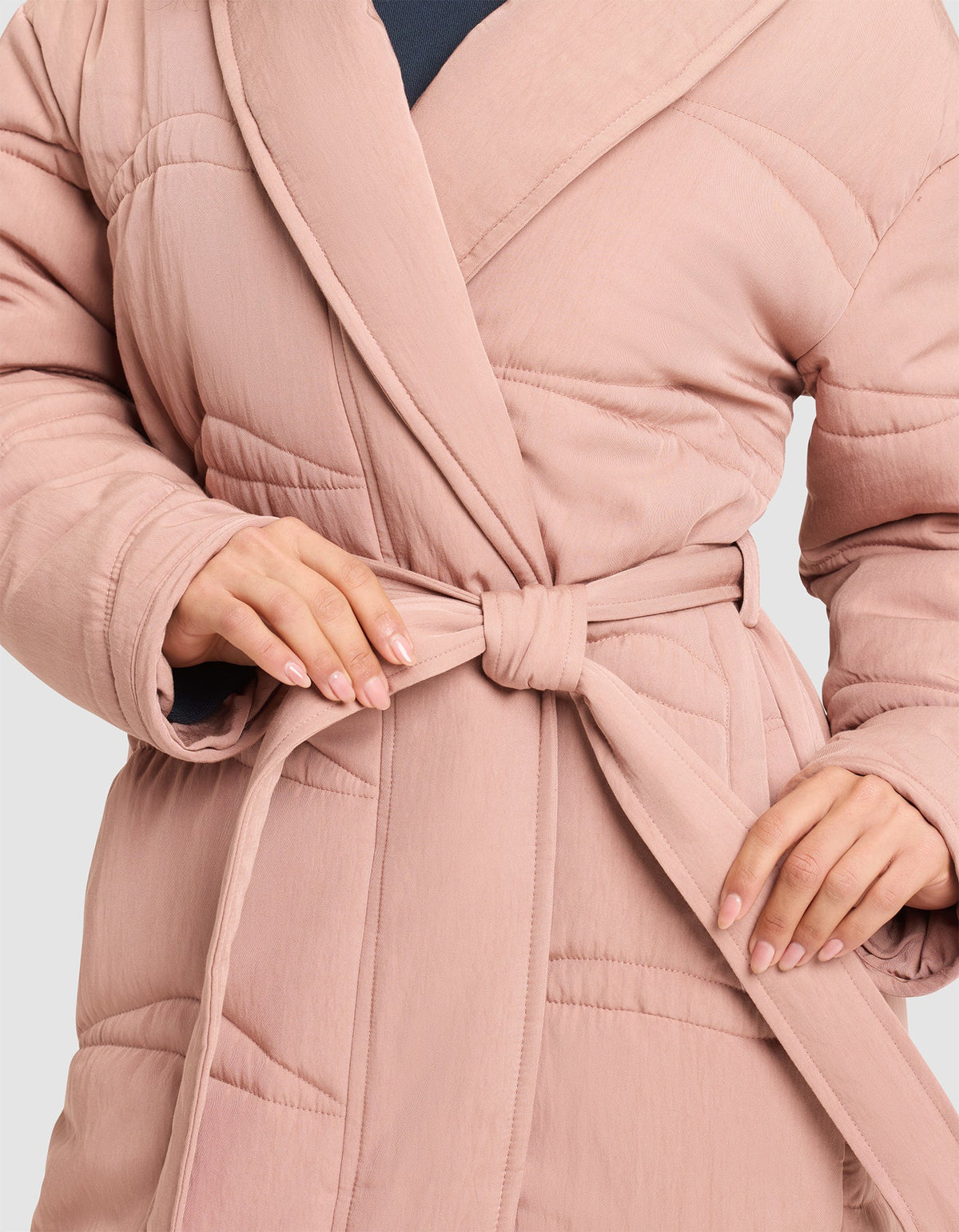 A person ties the waist belt of a Cozy Earth Women's Quilted House Coat in light pink, showing neatly manicured nails. The image highlights the hands and torso. |Color:Rosewood