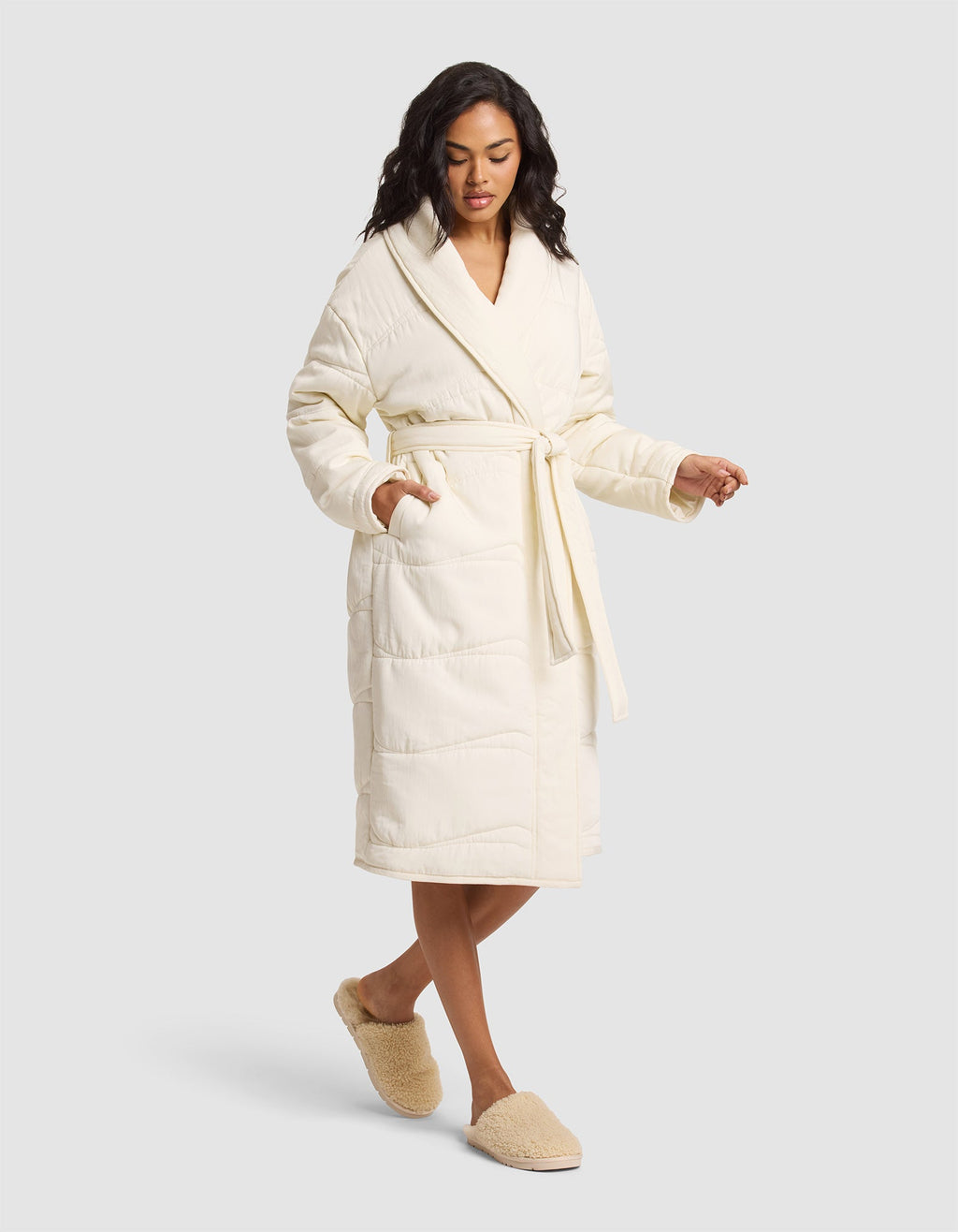 A woman models the Cozy Earth Women's Quilted House Coat, a long cream robe tied at the waist, paired with beige fuzzy slippers, standing against a plain light gray background. |Color:Vanilla