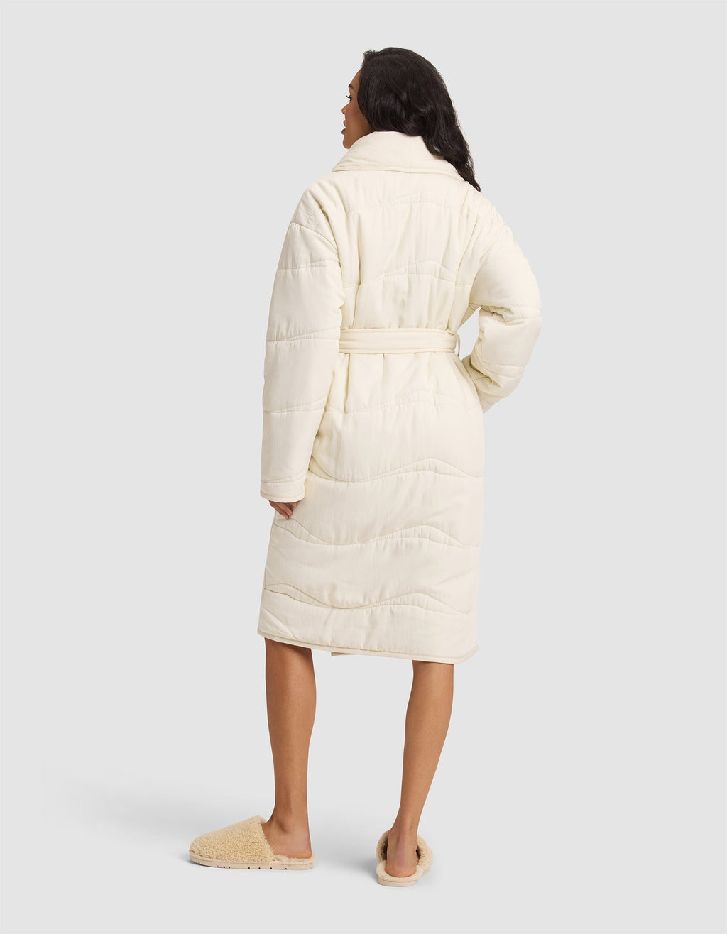 A woman faces away, wearing the Cozy Earth Women's Quilted House Coat in cream with a belt and tan slippers, set against a plain light gray background. |Color:Vanilla