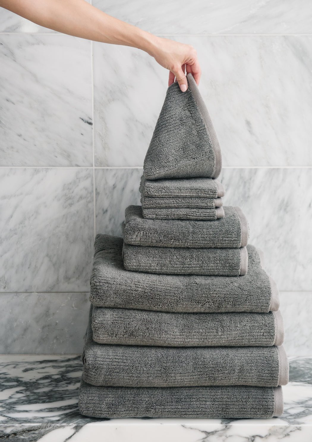 A hand places a folded gray Cozy Earth Complete Ribbed Terry Bath Bundle towel atop a neatly stacked pyramid of towels, set before a marble-tiled wall.|Color:Charcoal