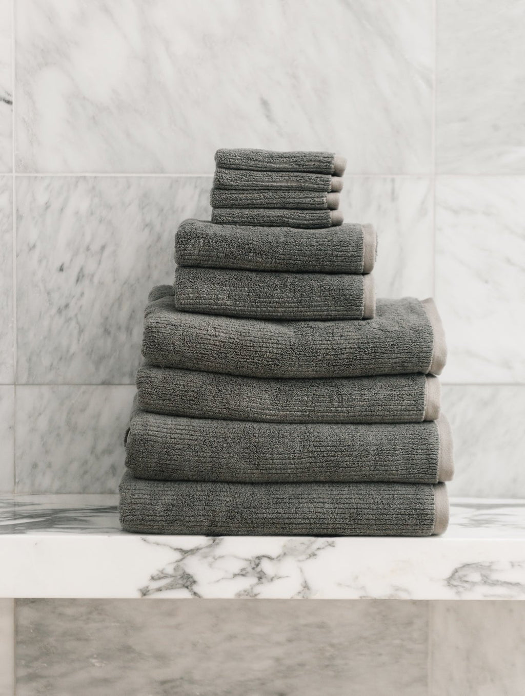 The Cozy Earth Complete Ribbed Terry Bath Bundle, featuring gray towels in assorted sizes, is displayed neatly on a white marble shelf with a marble-tiled wall backdrop.|Color:Charcoal
