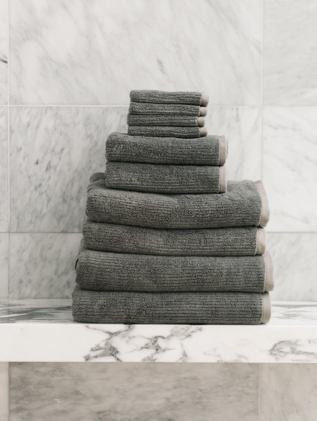 The Cozy Earth Complete Ribbed Terry Bath Bundle, featuring gray towels in assorted sizes, is displayed neatly on a white marble shelf with a marble-tiled wall backdrop.|Color:Charcoal