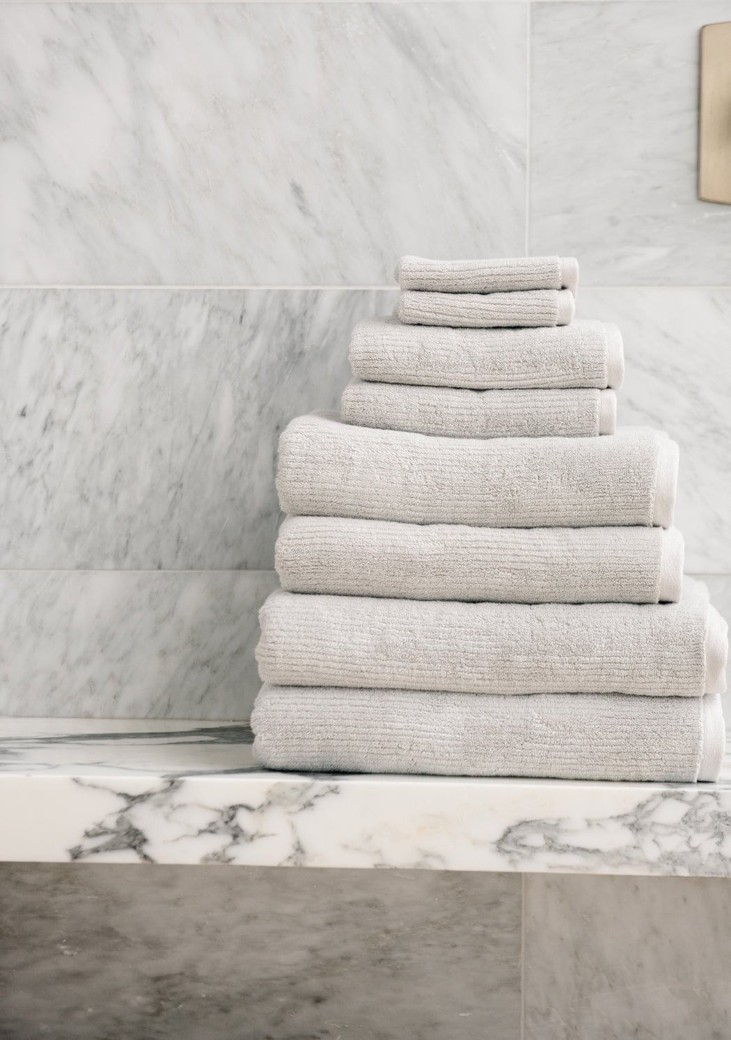 The Complete Ribbed Terry Bath Bundle by Cozy Earth is neatly stacked on a marble shelf in a bathroom with light gray marble walls.|Color:Light Grey