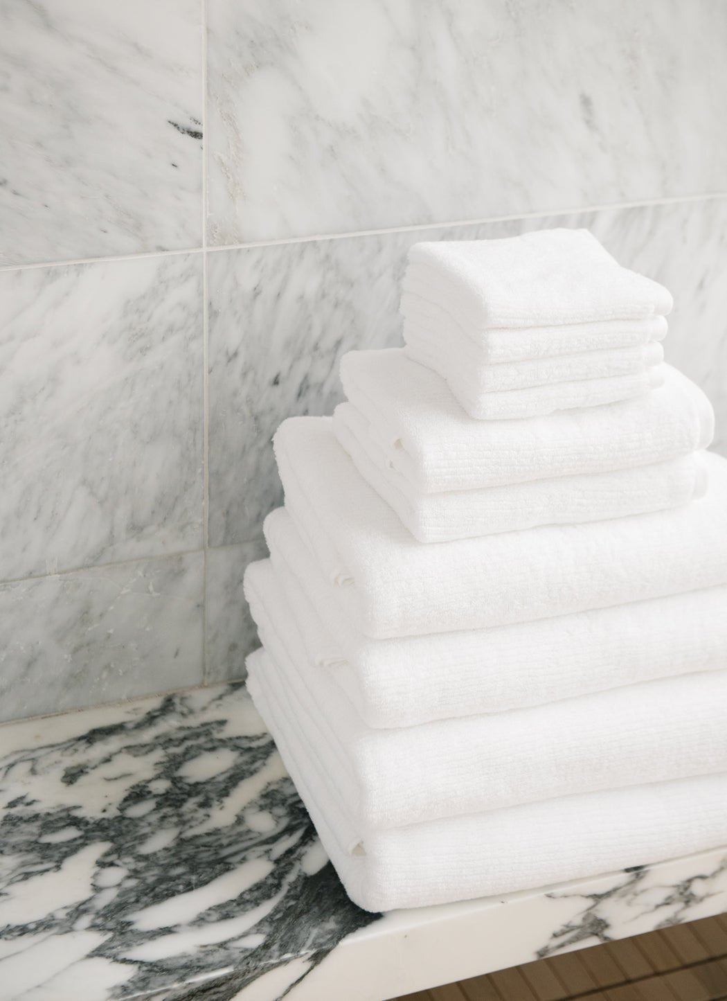 The Complete Ribbed Terry Bath Bundle by Cozy Earth is neatly stacked on a marble countertop, set against a gray and white marble wall.|Color:White