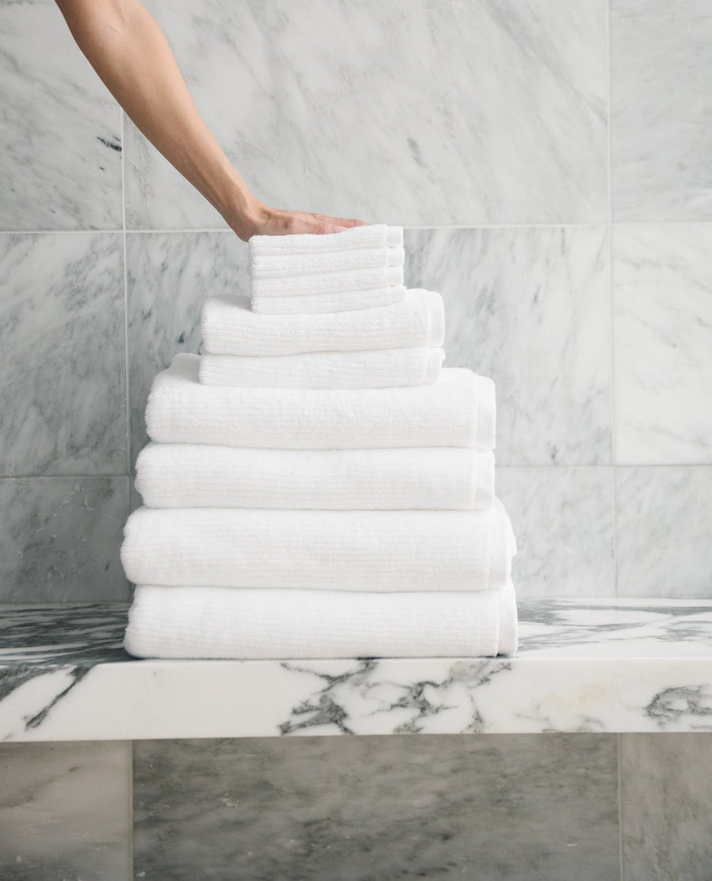 A hand places a neatly folded Cozy Earth Complete Ribbed Terry Bath Bundle towel atop a pyramid-shaped stack on a marble countertop, with a marble-tiled wall in the background.|Color:White