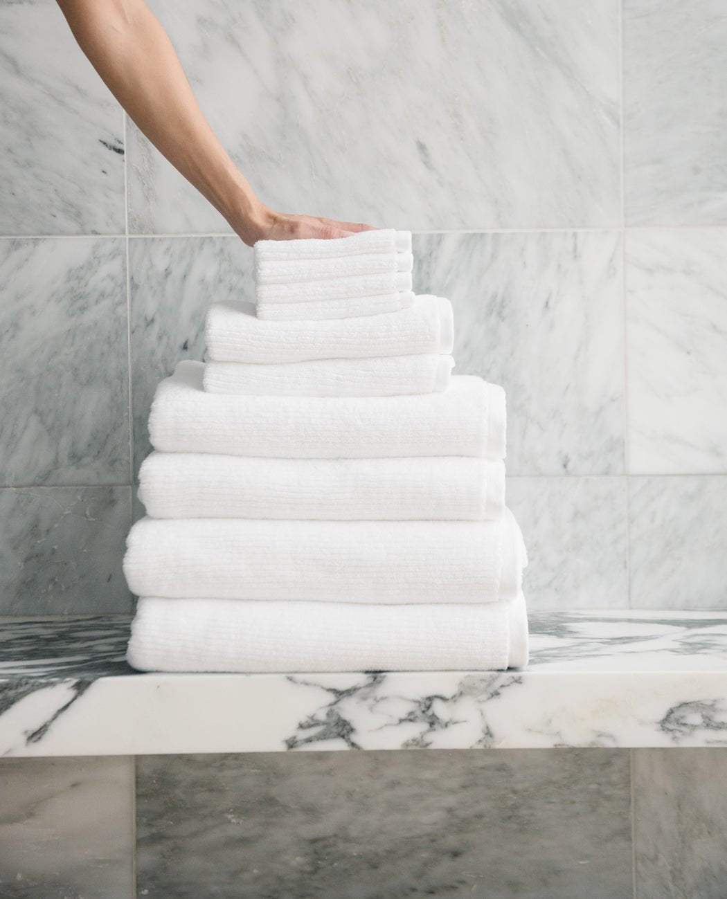 A hand places a neatly folded Cozy Earth Complete Ribbed Terry Bath Bundle towel atop a pyramid-shaped stack on a marble countertop, with a marble-tiled wall in the background.|Color:White