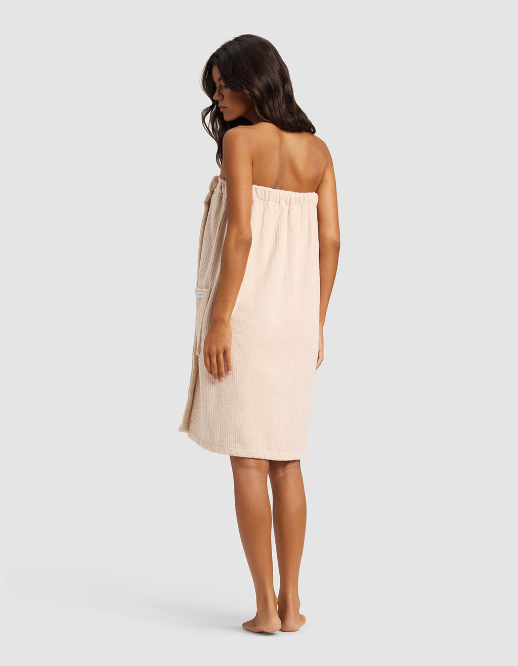A woman with long dark hair, barefoot and facing slightly away, wears the Cozy Earth Ribbed Terry Bath Wrap in beige against a plain light gray background.|Color:Petal