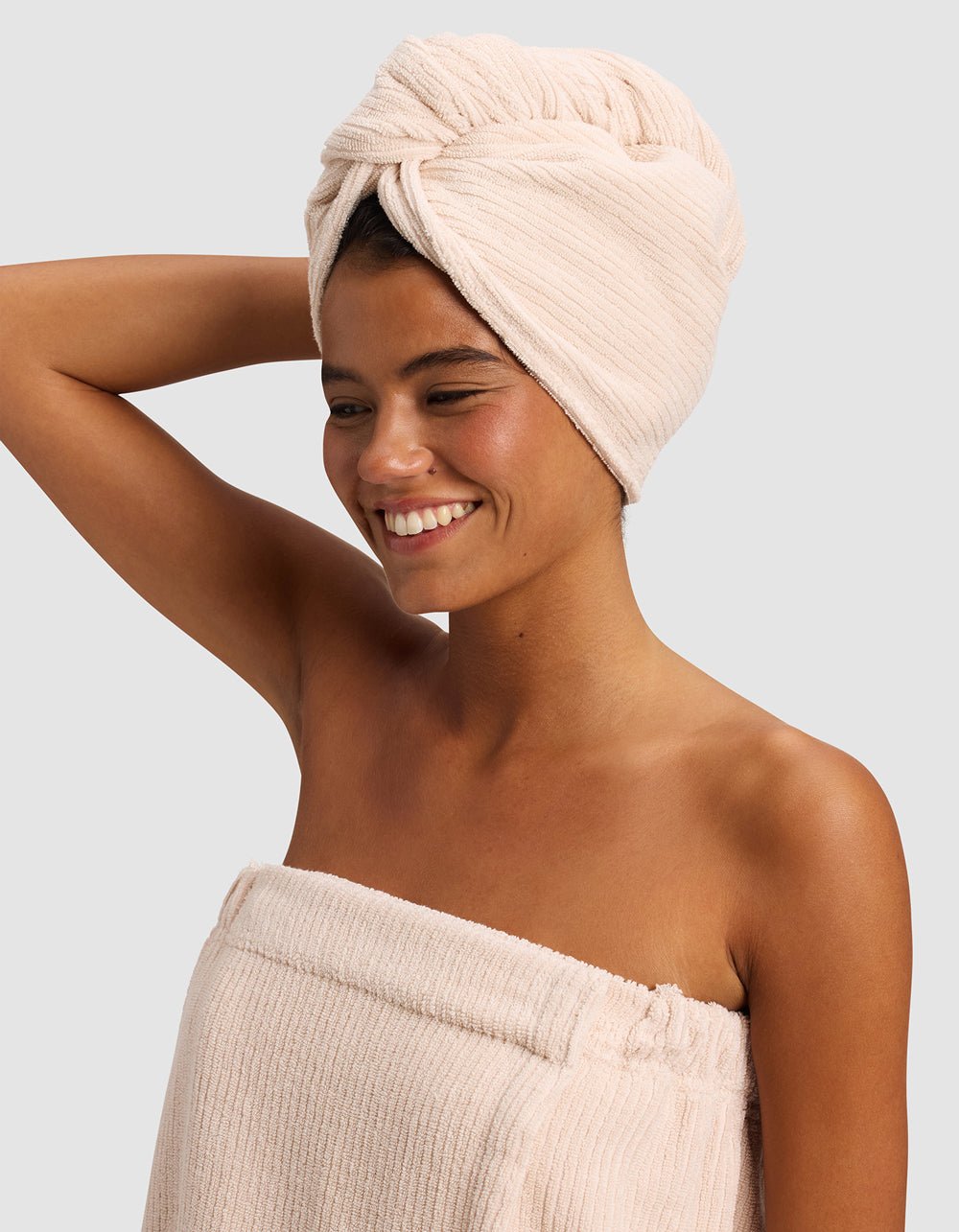 A smiling woman stands against a light gray background, wearing a beige towel wrap and the Cozy Earth Ribbed Terry Hair Towel in matching beige, with one arm raised and bent behind her head.|Color:Petal
