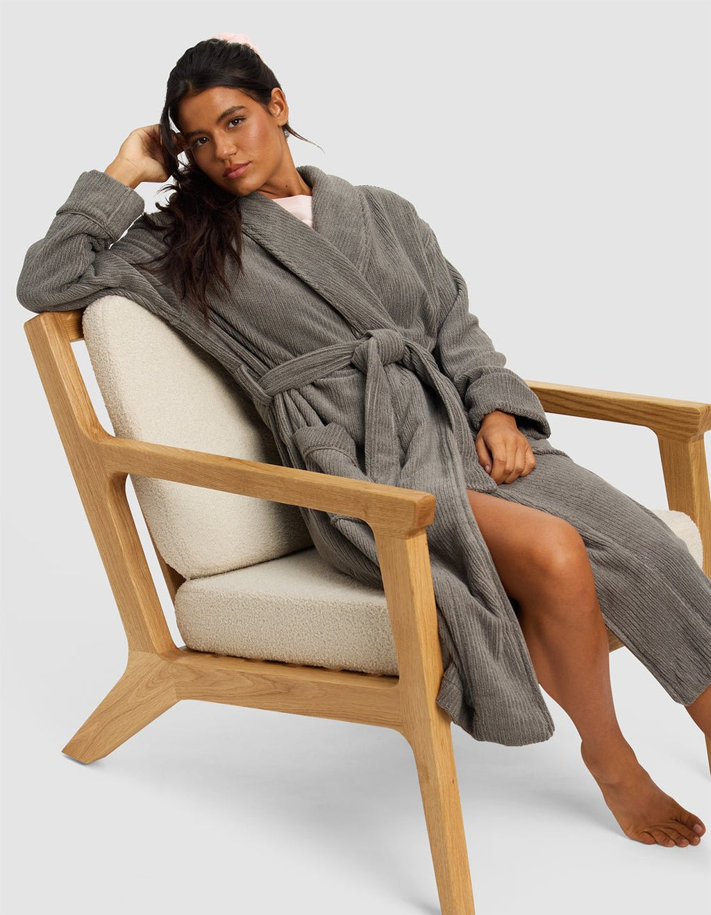 A woman with long dark hair sits barefoot on a modern wooden chair, wearing the Cozy Earth Ribbed Terry Bath Robe in gray. She rests her elbow on the armrest and gazes at the camera with a relaxed expression.|Color:Charcoal
