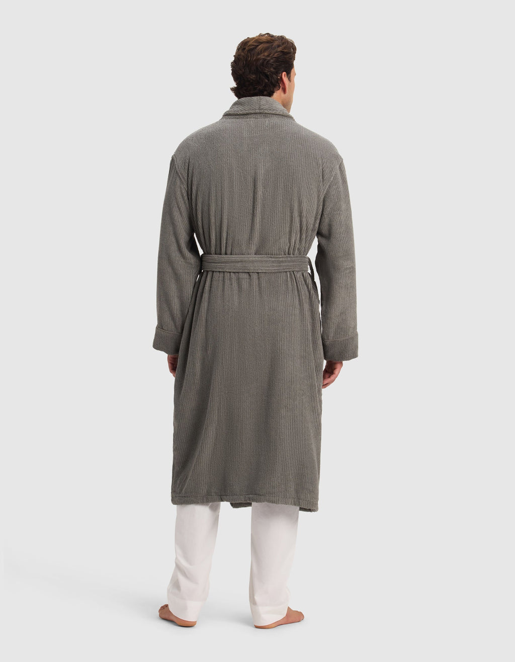A man, barefoot against a plain light background, is wearing the Cozy Earth Ribbed Terry Bath Robe in gray, featuring a textured finish and waist belt, styled over white pants.|Color:Charcoal
