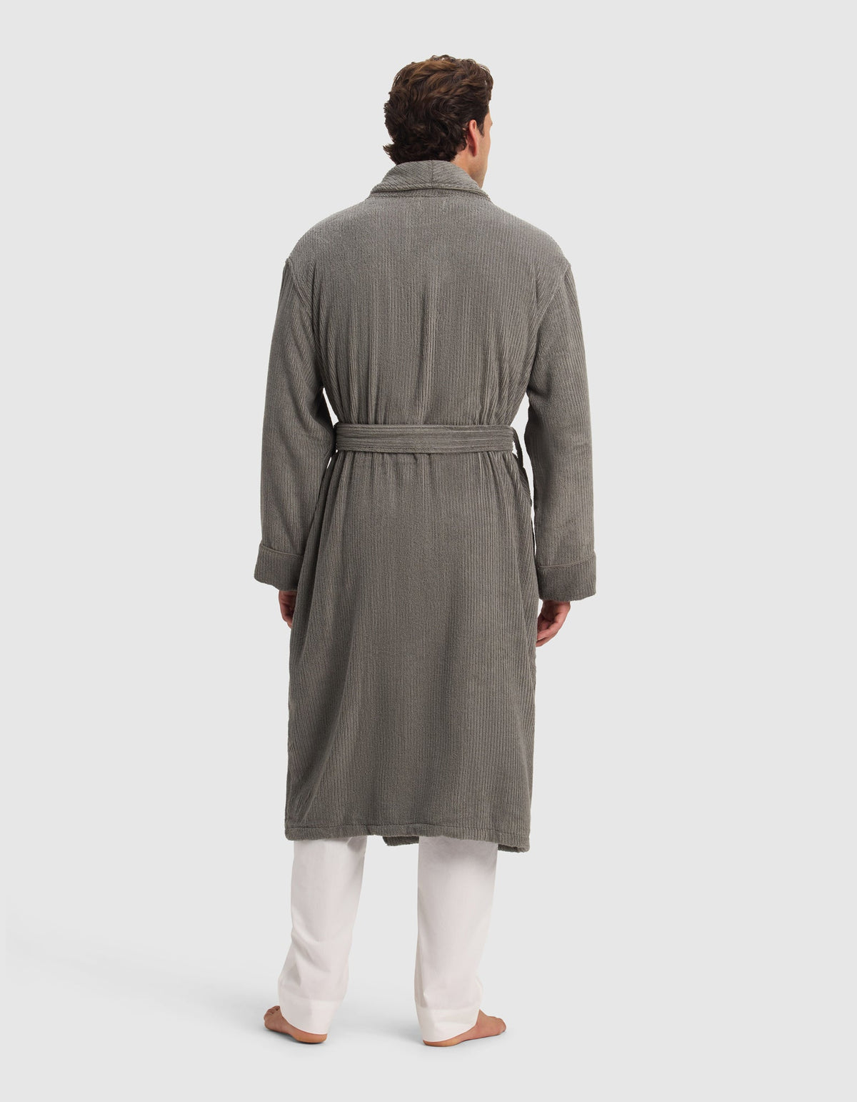 A man, barefoot against a plain light background, is wearing the Cozy Earth Ribbed Terry Bath Robe in gray, featuring a textured finish and waist belt, styled over white pants.|Color:Charcoal
