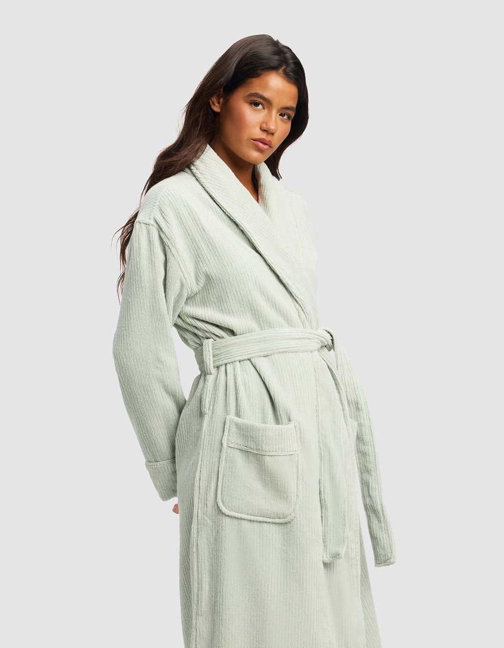 A woman with long dark hair wears the Cozy Earth Ribbed Terry Bath Robe in light sage green, featuring a belted waist and large front pockets, standing against a plain light gray background.|Color:Laurel