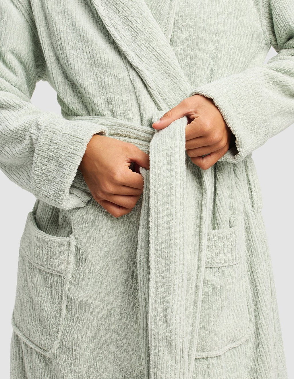 A person ties the belt of a Cozy Earth Ribbed Terry Bath Robe in light green. The textured robe features large front pockets and visible ribbing, with their hands adjusting the waist for comfort.|Color:Laurel