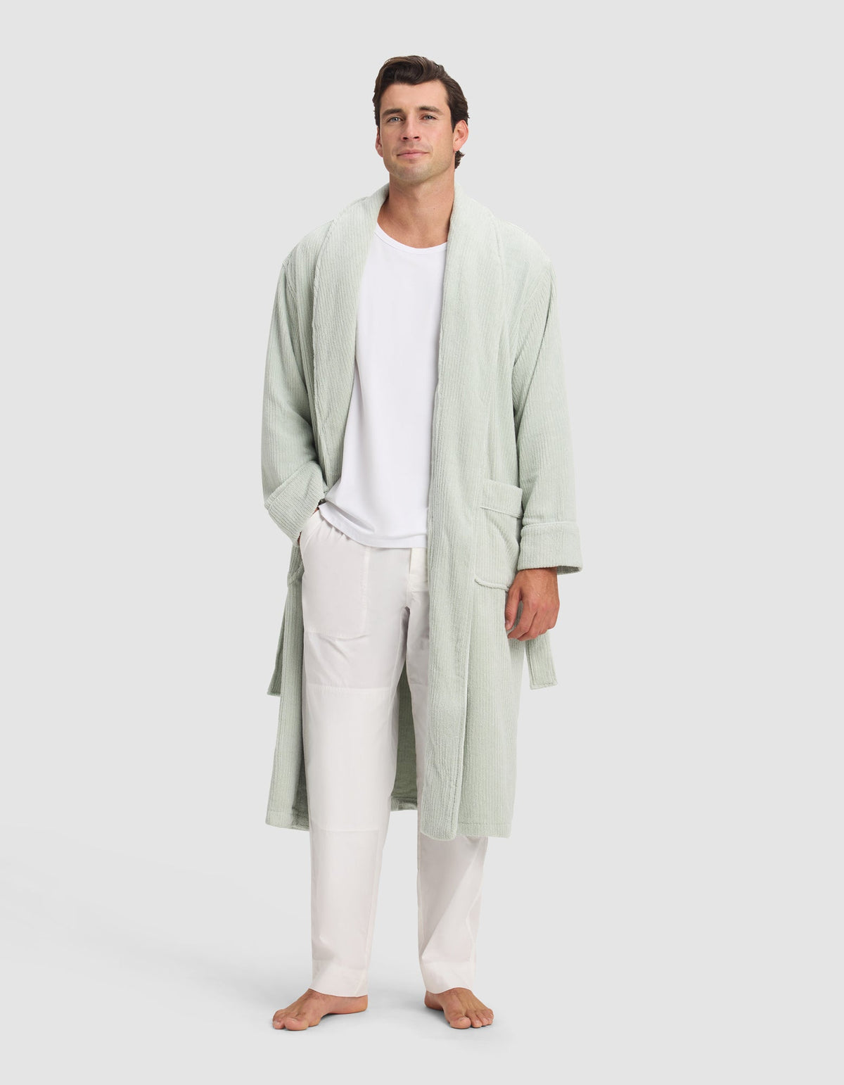 A man stands barefoot against a light background, wearing the Cozy Earth Ribbed Terry Bath Robe in light green over a white T-shirt and white pants, with his hands in the robe's pockets.|Color:Laurel