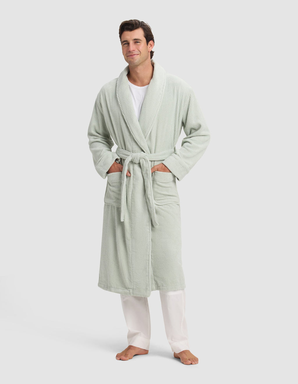 A man smiles barefoot in a Cozy Earth Ribbed Terry Bath Robe—light green with a belt and two pockets—styled over a white shirt and pants, set against a plain light background.|Color:Laurel