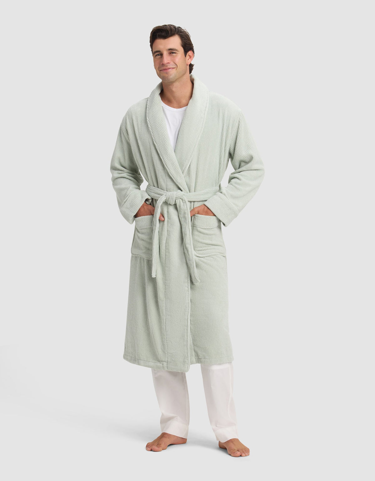 A man smiles barefoot in a Cozy Earth Ribbed Terry Bath Robe—light green with a belt and two pockets—styled over a white shirt and pants, set against a plain light background.|Color:Laurel