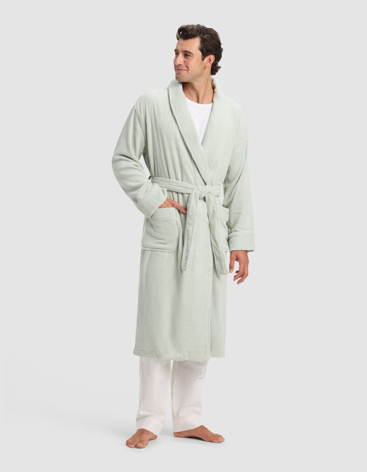 A man stands barefoot, wearing Cozy Earth's Ribbed Terry Bath Robe in light green, belted over a white shirt and pants. One hand is in his pocket as he looks to the side against a plain, light background.|Color:Laurel
