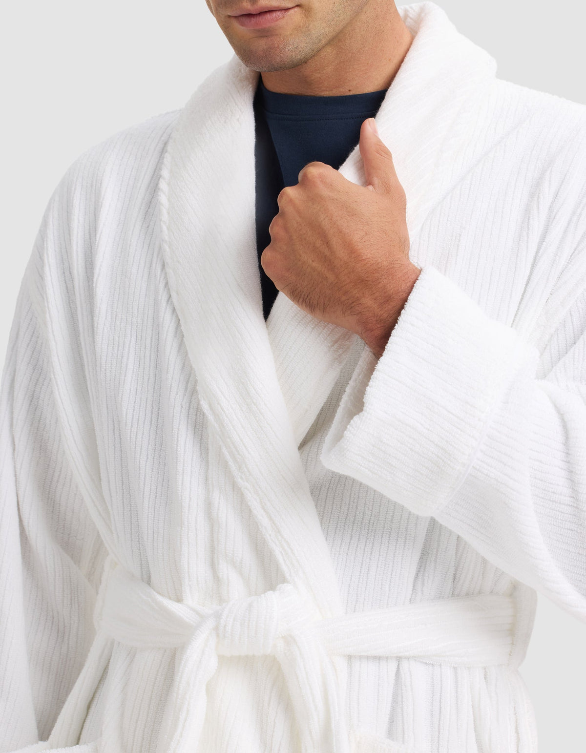 Someone wearing the Cozy Earth Ribbed Terry Bath Robe in white over a dark shirt, tying the belt at the waist and holding the collar with one hand. The person's face is partly visible.|Color:White