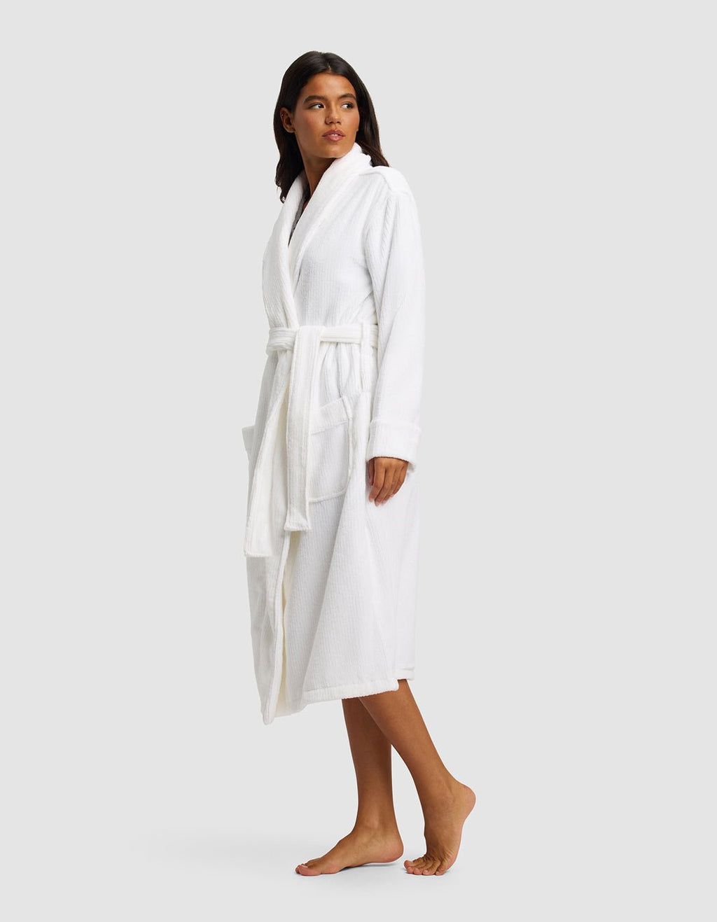 A woman stands barefoot, wearing the Cozy Earth Ribbed Terry Bath Robe—a white, knee-length robe with a belt and large front pockets—looking slightly left against a plain light gray background.|Color:White