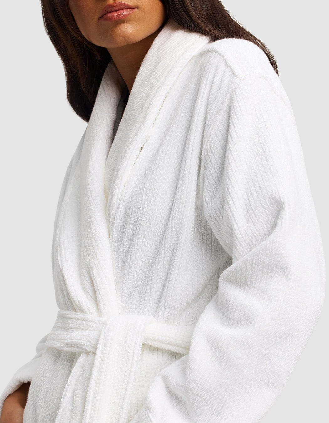 A woman models the Cozy Earth Ribbed Terry Bath Robe, featuring a plush white fabric, shawl collar, and belt. She stands against a plain light background with only her lower face and upper body visible.|Color:White