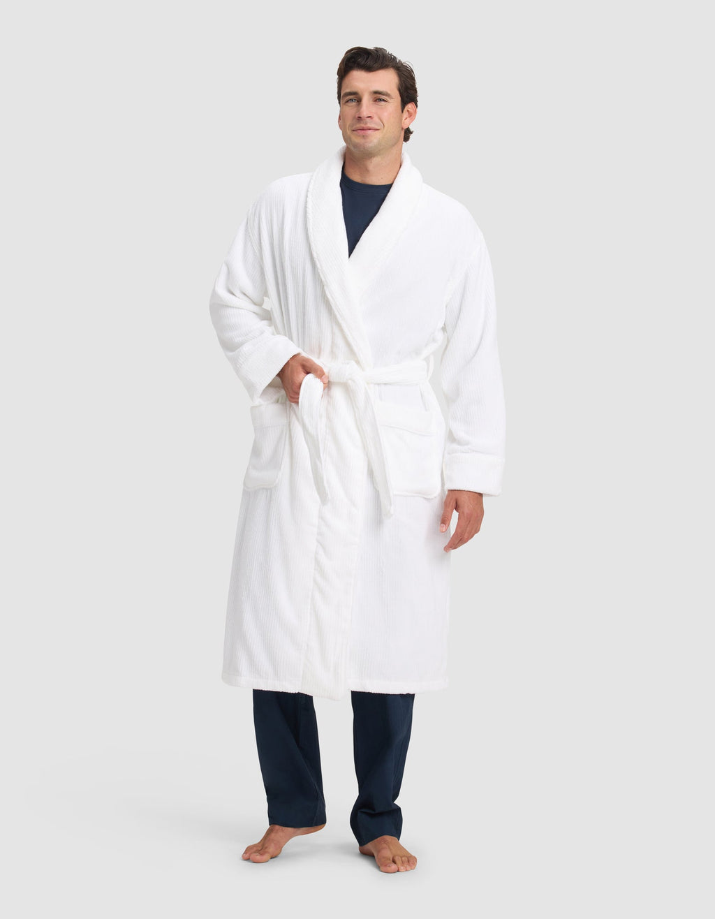 A person stands barefoot in a Cozy Earth Ribbed Terry Bath Robe over dark blue pajamas, posing against a light background with one hand in a pocket and the other at their side.|Color:White