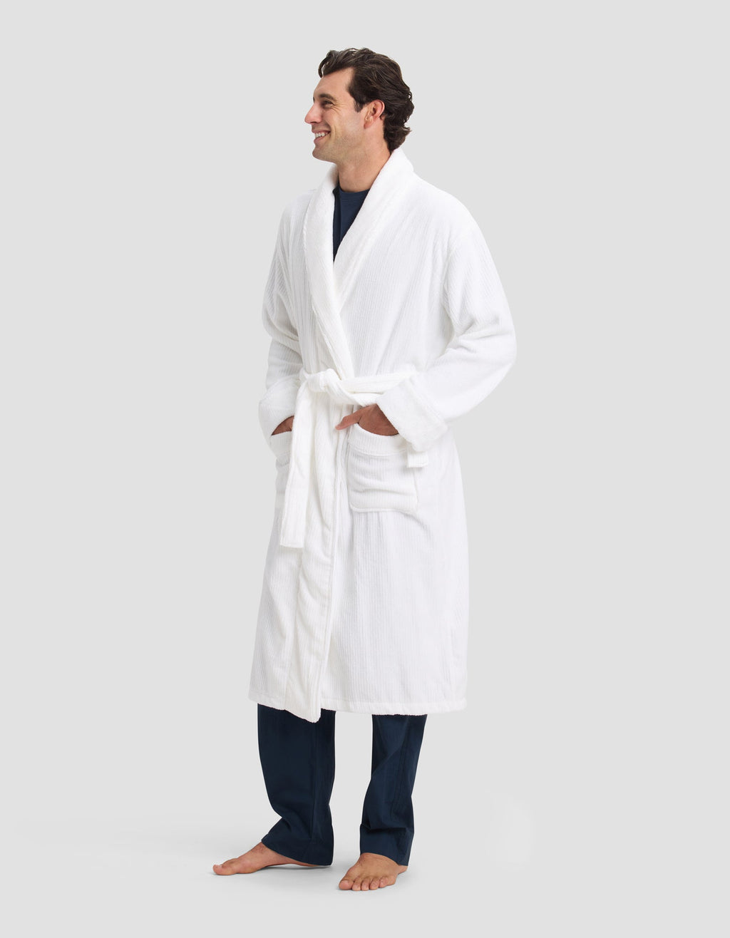 A man stands barefoot, smiling, with his hands in the pockets of a Cozy Earth Ribbed Terry Bath Robe tied at the waist over navy pants and a navy shirt. The background is plain light gray.|Color:White