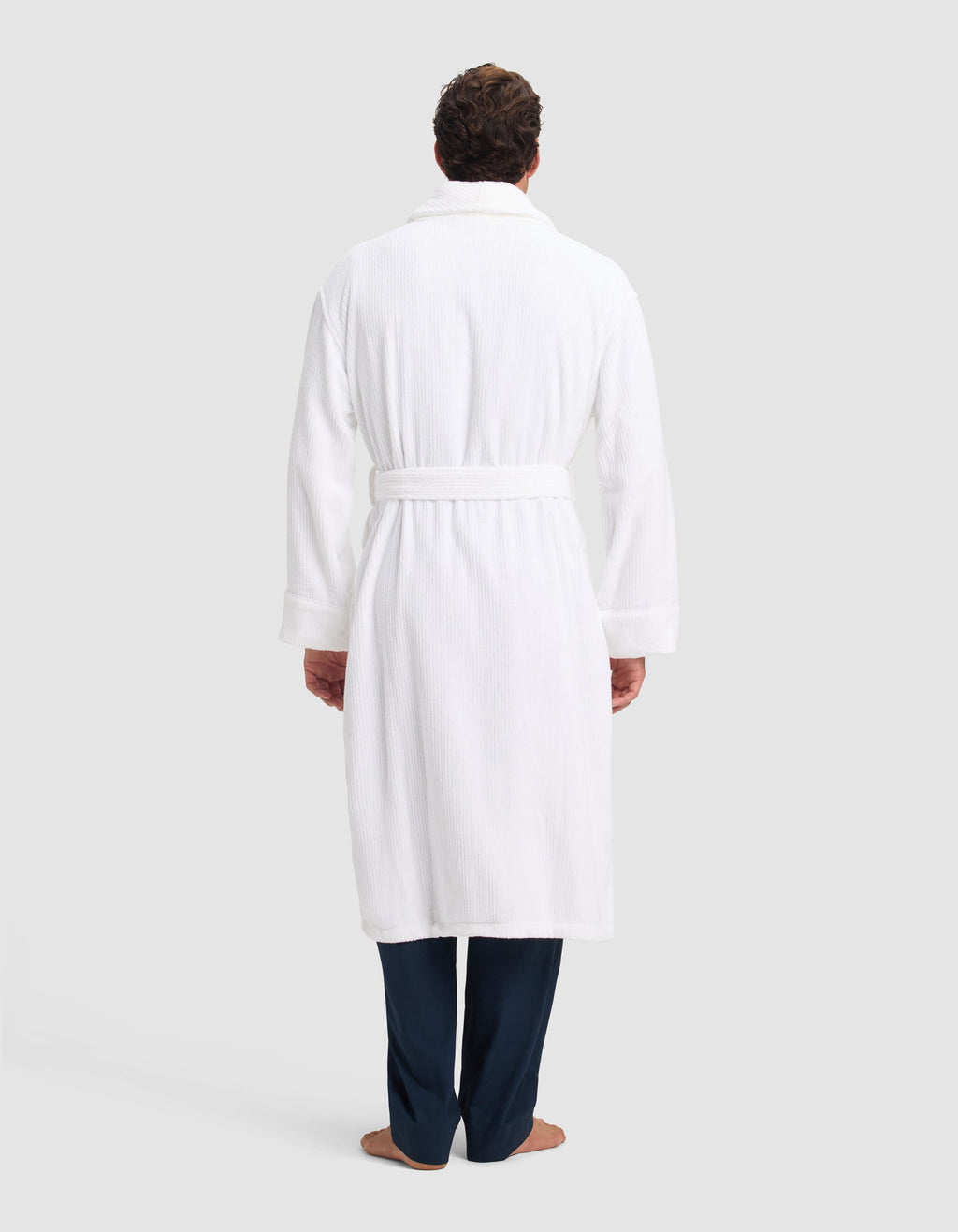 A person faces away, wearing the Cozy Earth Ribbed Terry Bath Robe with a belt, dark pants, and bare feet, set against a plain light background.|Color:White