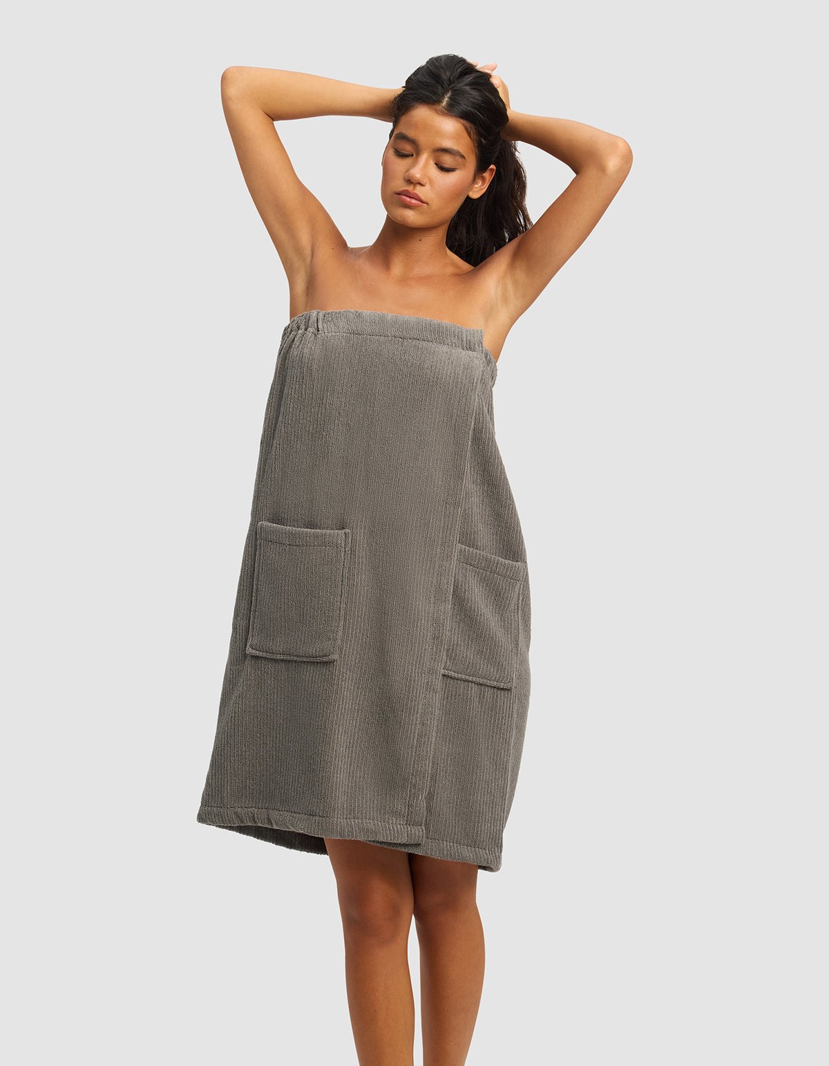 A woman stands with her hands behind her head, wearing the gray Ribbed Terry Bath Wrap by Cozy Earth, featuring a strapless design and two front pockets. The background is plain and light-colored. |Color:Charcoal