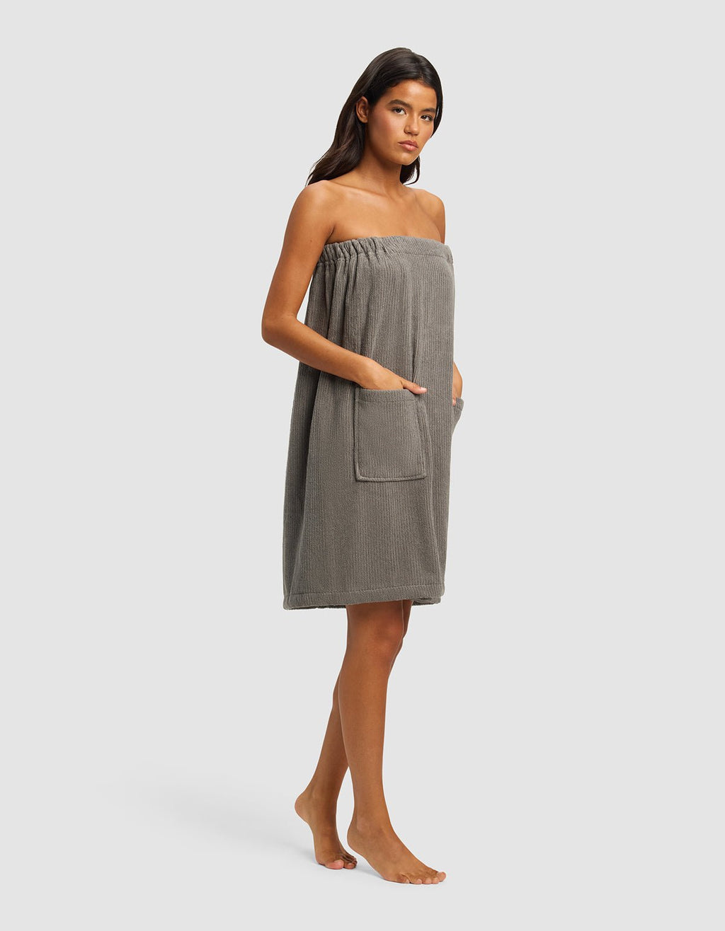 A woman stands barefoot against a plain background, wearing the Cozy Earth Ribbed Terry Bath Wrap in grey. Its strapless design features pockets, and she rests her hands in them. Her straight dark hair frames her neutral expression.|Color:Charcoal