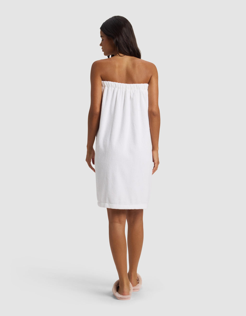 A woman with long dark hair stands facing away, wearing the Cozy Earth Ribbed Terry Bath Wrap and pink slippers against a plain light background.|Color:White