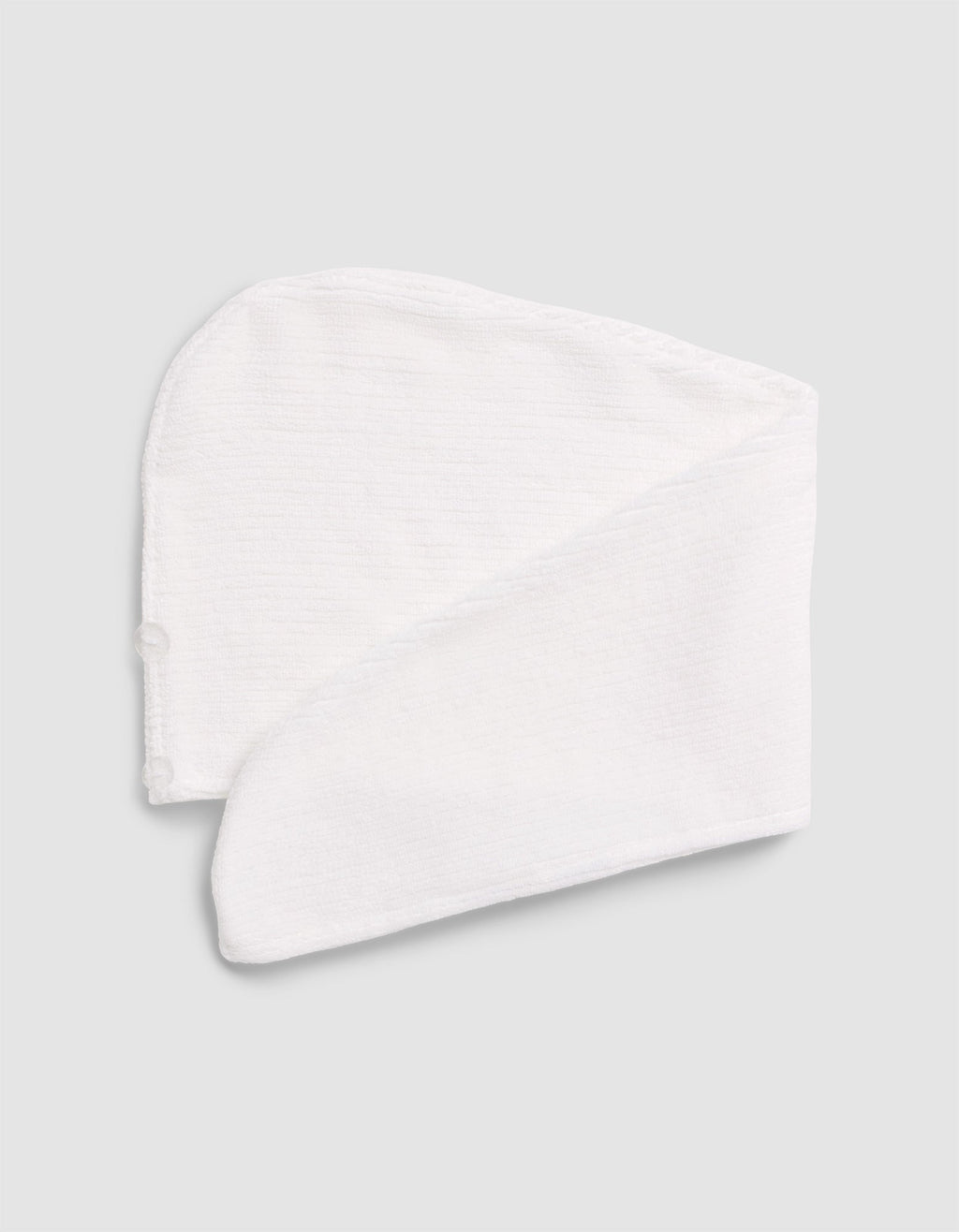 The Cozy Earth Ribbed Terry Hair Towel, in white textured fabric, is folded flat on a light gray surface. It features a curved top, long tapered end, and a small side button.|Color:White