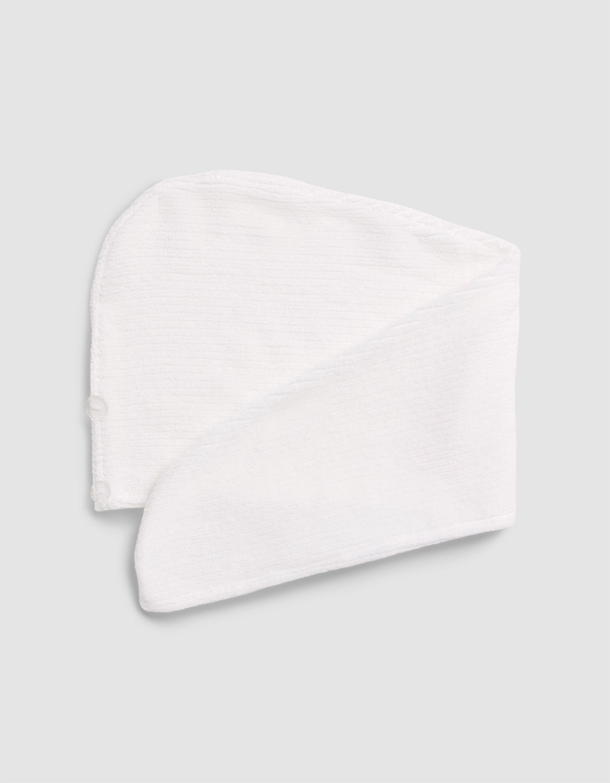 The Cozy Earth Ribbed Terry Hair Towel, in white textured fabric, is folded flat on a light gray surface. It features a curved top, long tapered end, and a small side button.|Color:White