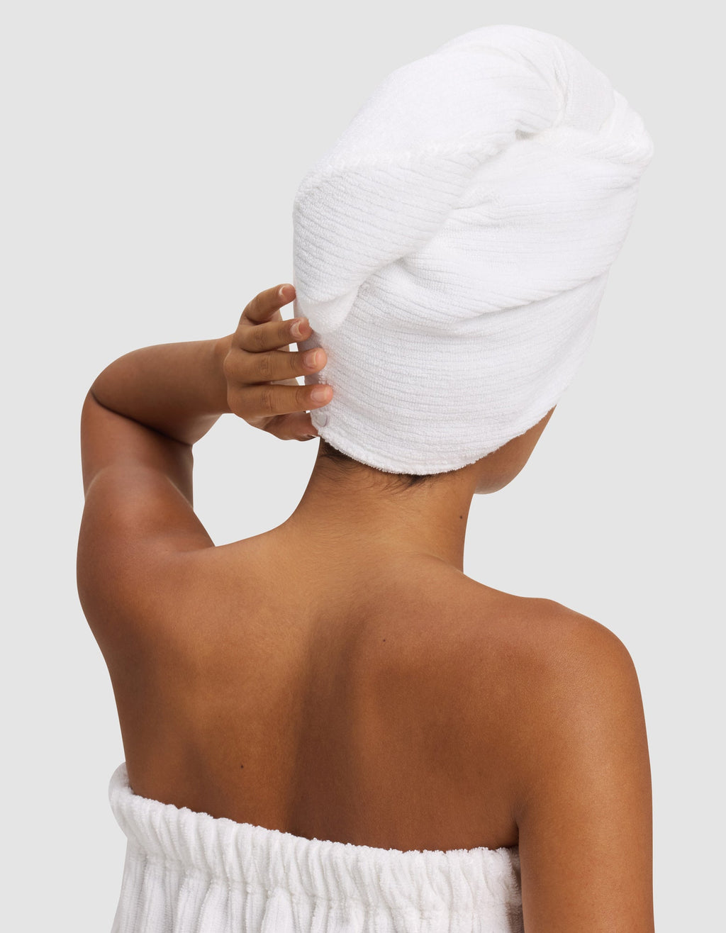 A person with medium skin tone is seen from behind, wearing a white towel around their body and holding a Cozy Earth Ribbed Terry Hair Towel wrapped around their head. The background is plain light gray.|Color:White