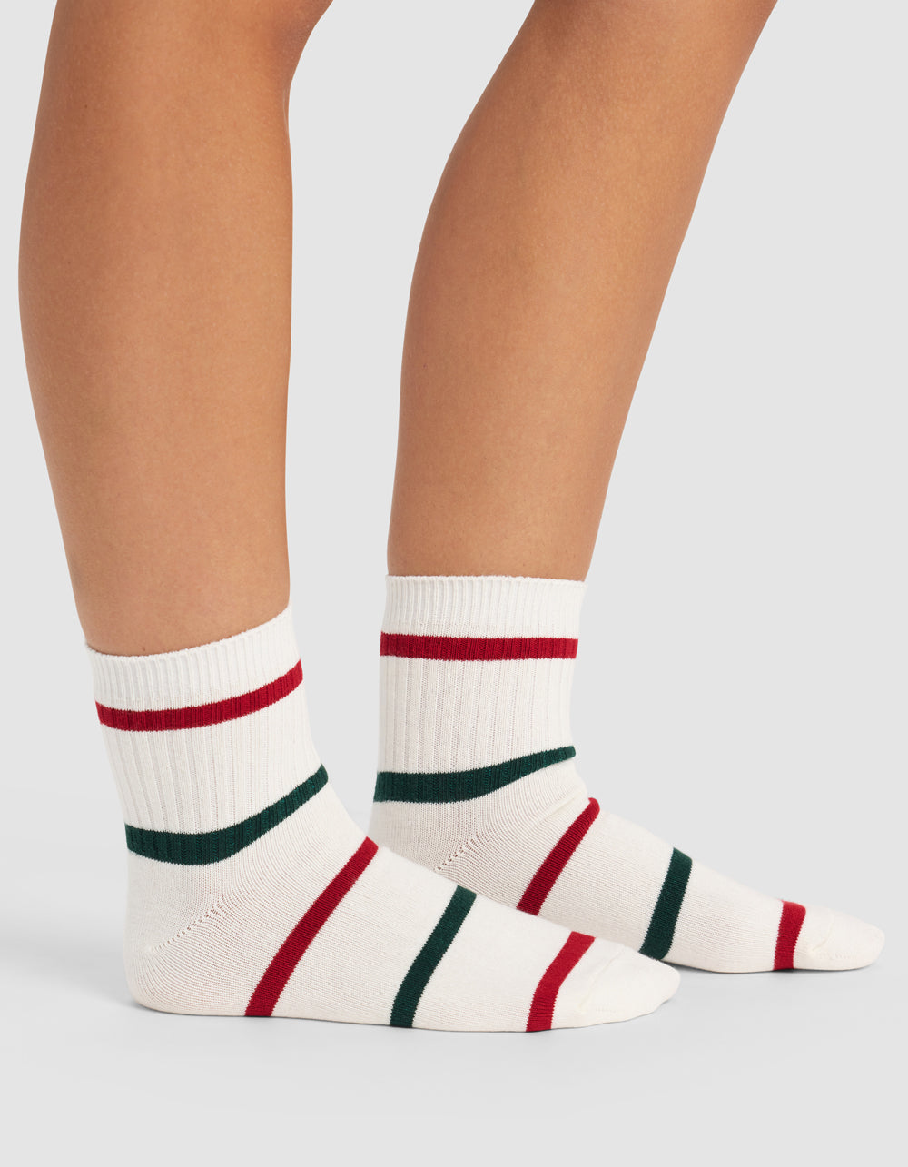 A person wears Cozy Earth Women's Sadie Socks—white ribbed crew socks with red and green stripes—standing on a light gray surface, showing only their lower legs and feet.|Color:Jade/Crimson