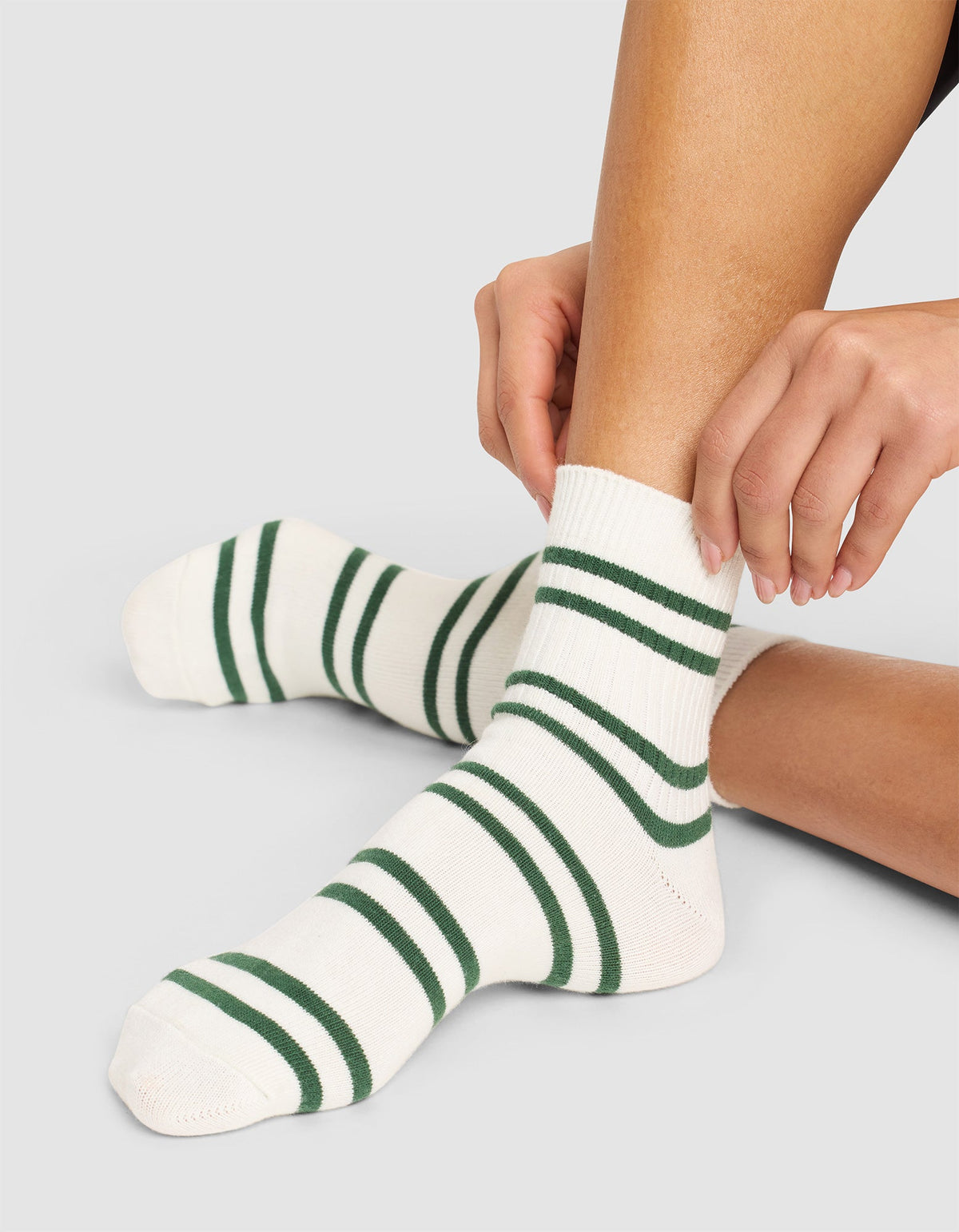 A person wearing Cozy Earth Women's Sadie Socks in white with dark green stripes sits on the floor, adjusting one sock. The photo highlights their legs and feet. |Color:Celadon/Jade