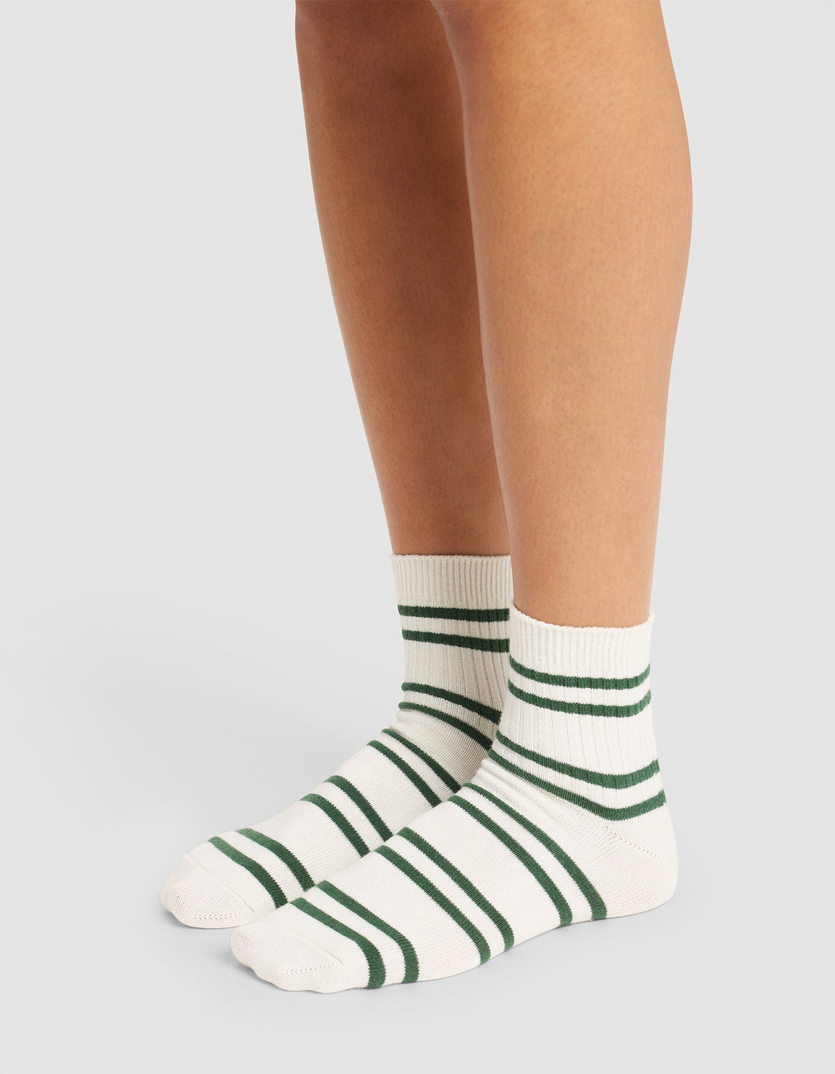 A person wears Cozy Earth Women's Sadie Socks—white with green horizontal stripes reaching just above the ankles—while standing on a light-colored surface. Only their legs and feet are visible. |Color:Celadon/Jade