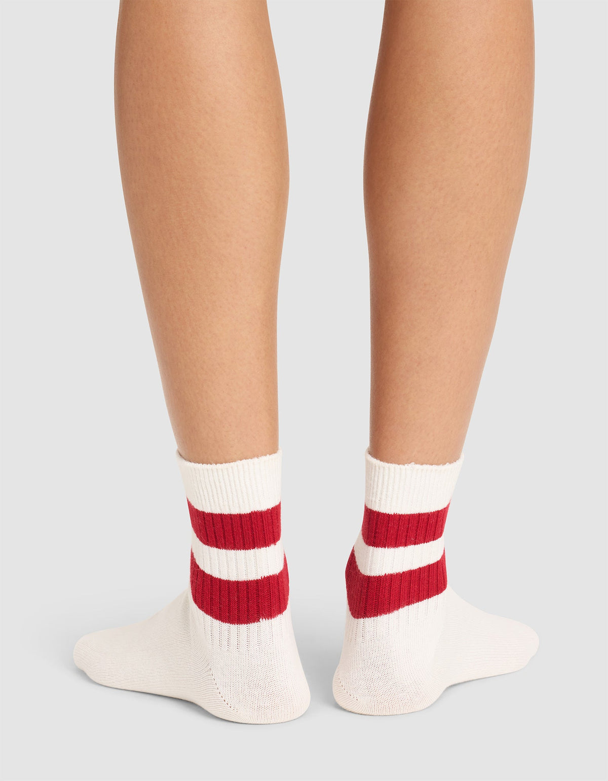 Back view of a person wearing Cozy Earth Women's Sadie Socks—white with two thick red stripes—standing on a light gray surface. Only the lower legs and feet are shown.  |Color:Bone/Crimson