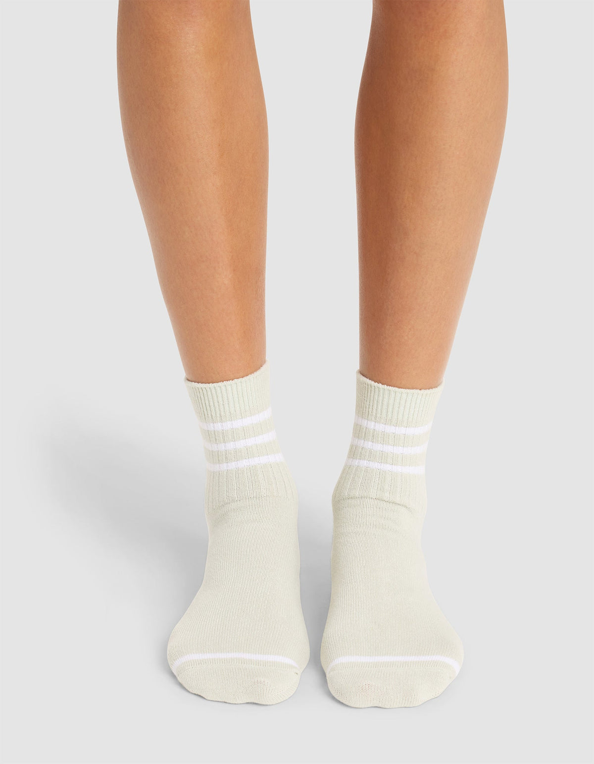 A person wearing Cozy Earth's Women's Sadie Sock in off-white with three white cuff stripes stands on a light grey surface, showing only their lower legs and feet. |Color:Laurel/White