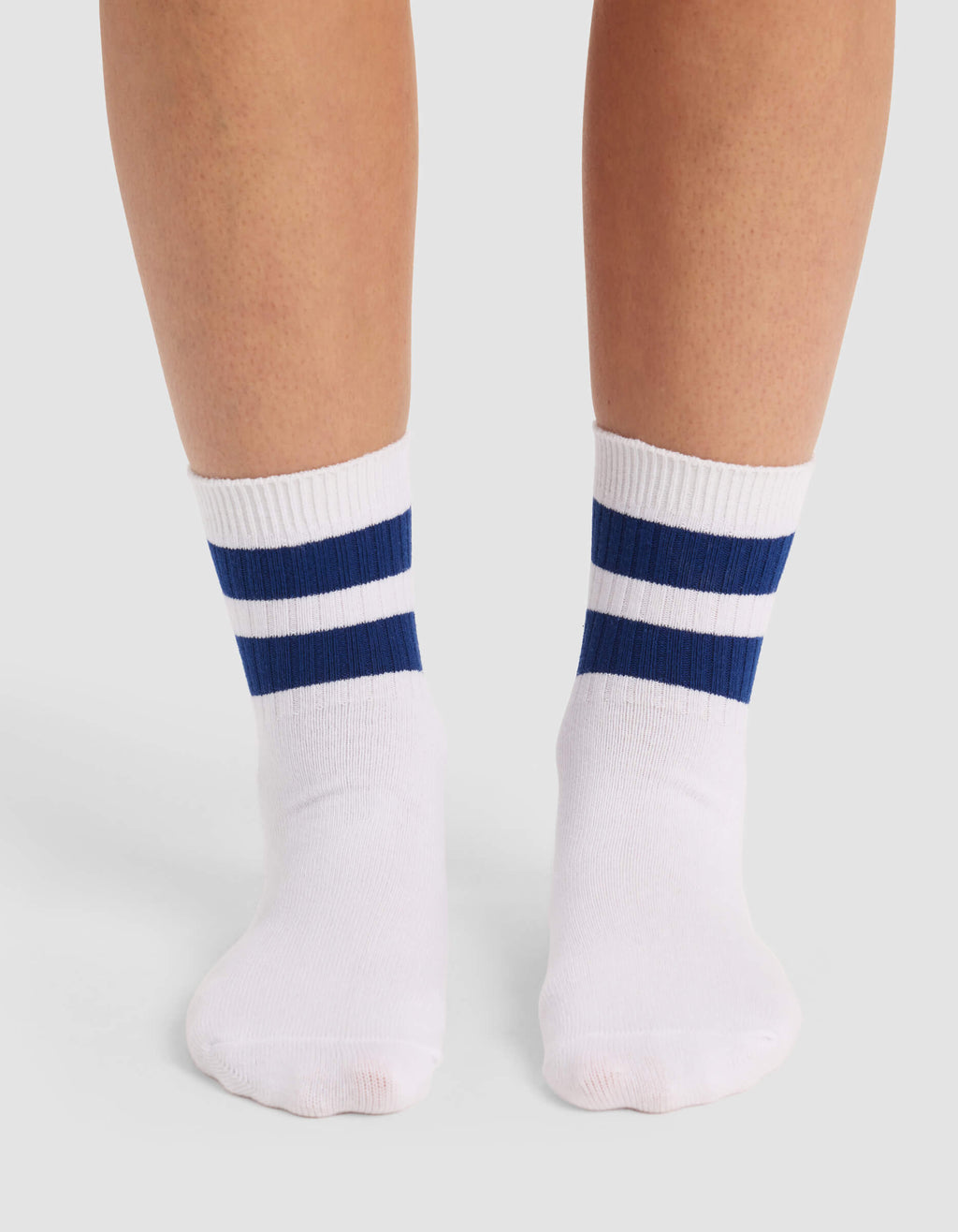 A person models Cozy Earth's Women's Sadie Sock, featuring two navy blue stripes near the top, while standing on a light gray surface.|Color:White/Pacific