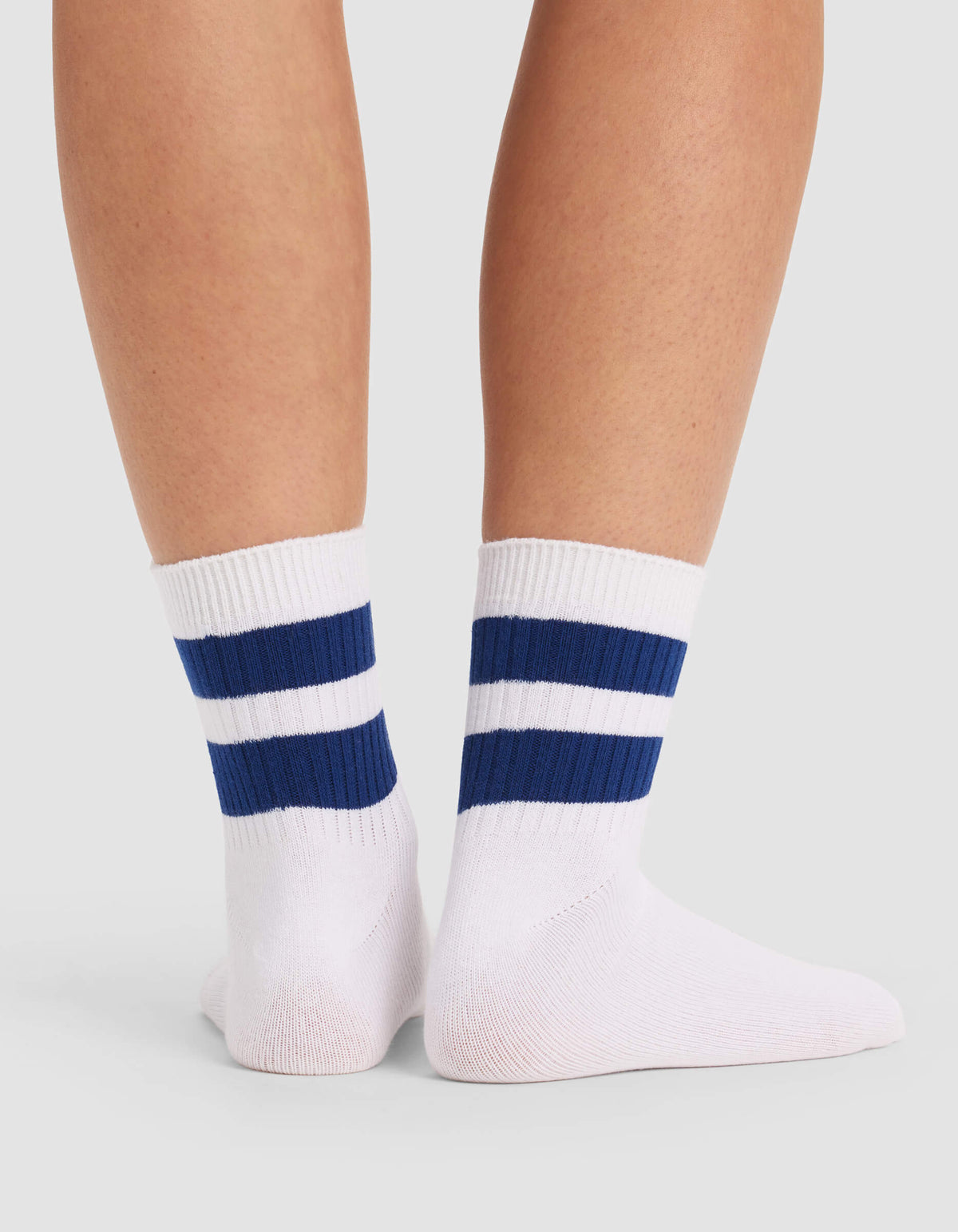 A pair of feet wearing Cozy Earth Women's Sadie Socks—white crew socks with two thick blue stripes near the top—standing against a light gray background.|Color:White/Pacific