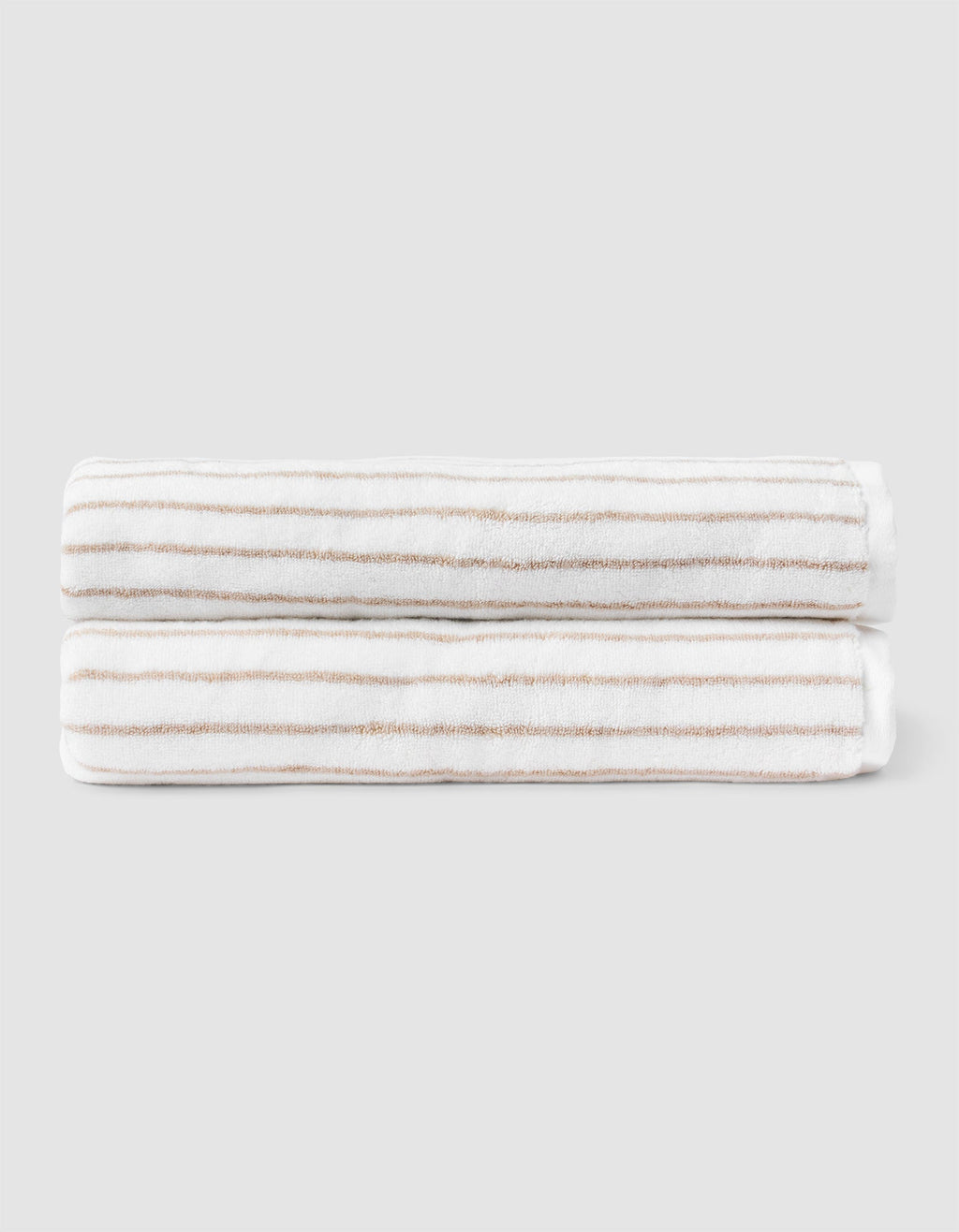 Two Striped Luxe Bath Sheets by Cozy Earth, featuring white fabric with thin, evenly spaced tan stripes, are neatly folded and stacked against a plain light gray background.  |Color:Sand Stripe