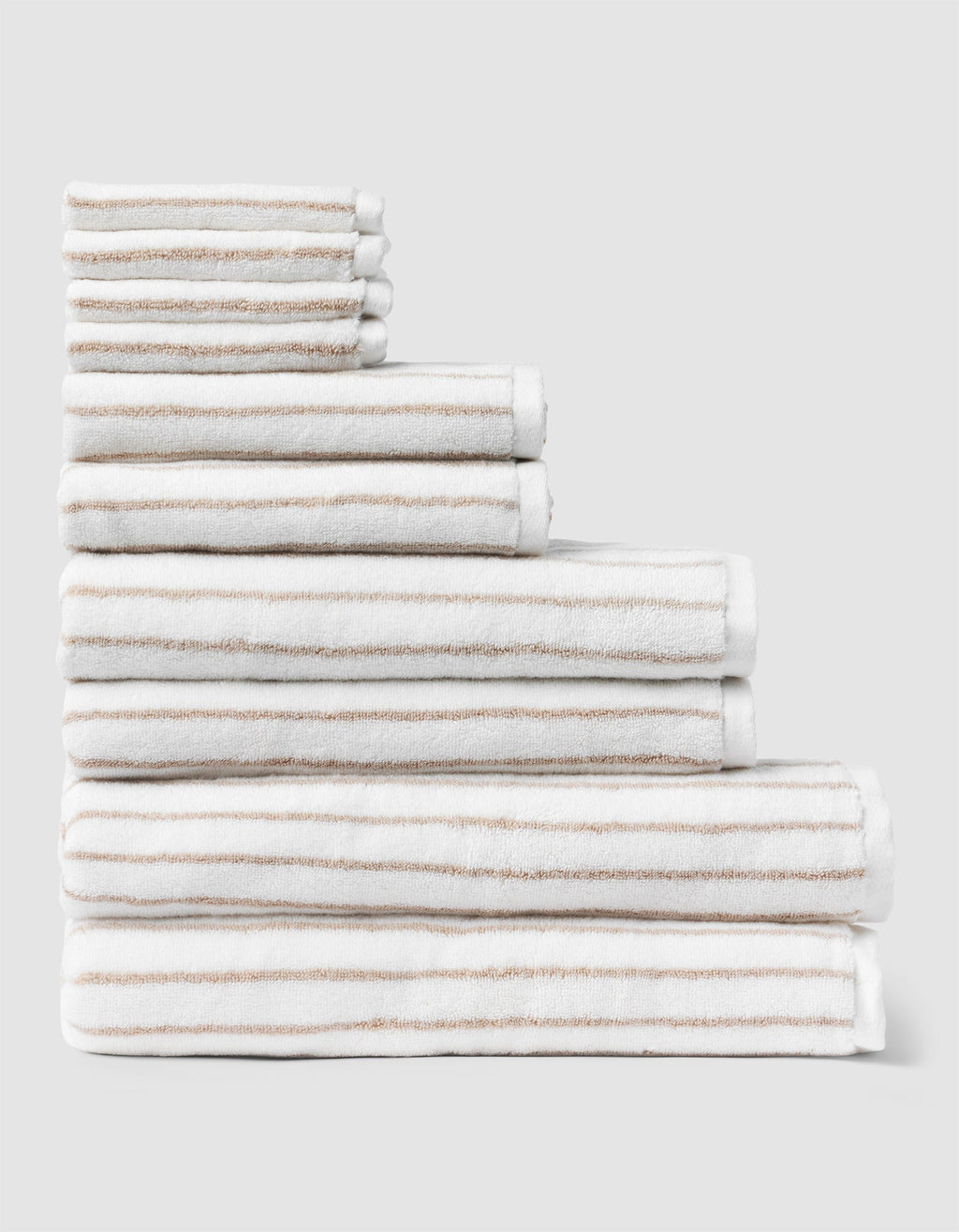 The Complete Striped Luxe Bath Bundle by Cozy Earth features neatly stacked white towels with thin beige stripes, arranged in descending size order against a plain light gray background. |Color:Sand Stripe