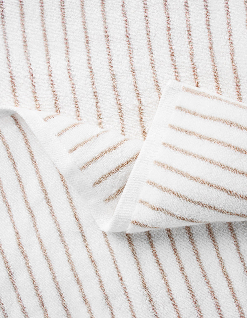 A close-up of the Cozy Earth Striped Luxe Hand Towels, featuring thin brown vertical stripes on white fabric, shown partially folded with one corner overlapping. |Color:Sand Stripe