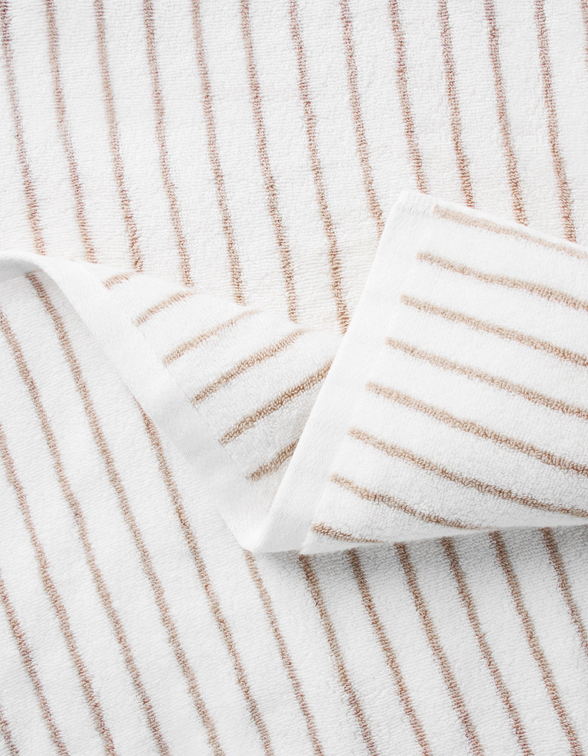 A close-up of the Cozy Earth Striped Luxe Hand Towels, featuring thin brown vertical stripes on white fabric, shown partially folded with one corner overlapping. |Color:Sand Stripe