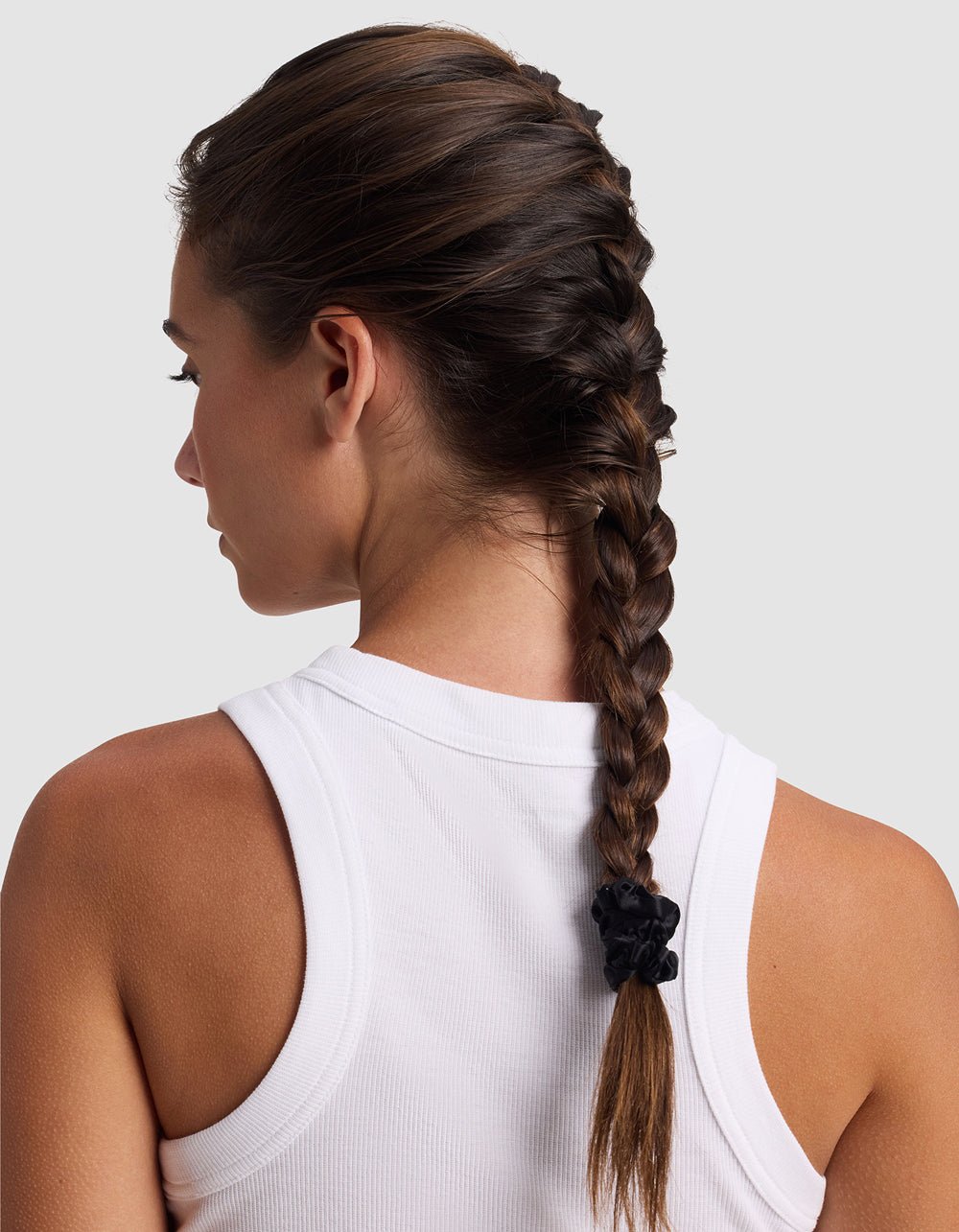 A woman with brown hair in a French braid, secured with a Cozy Earth Silk Skinny Scrunchie, wears a white sleeveless top and is seen from the back against a light gray background.
