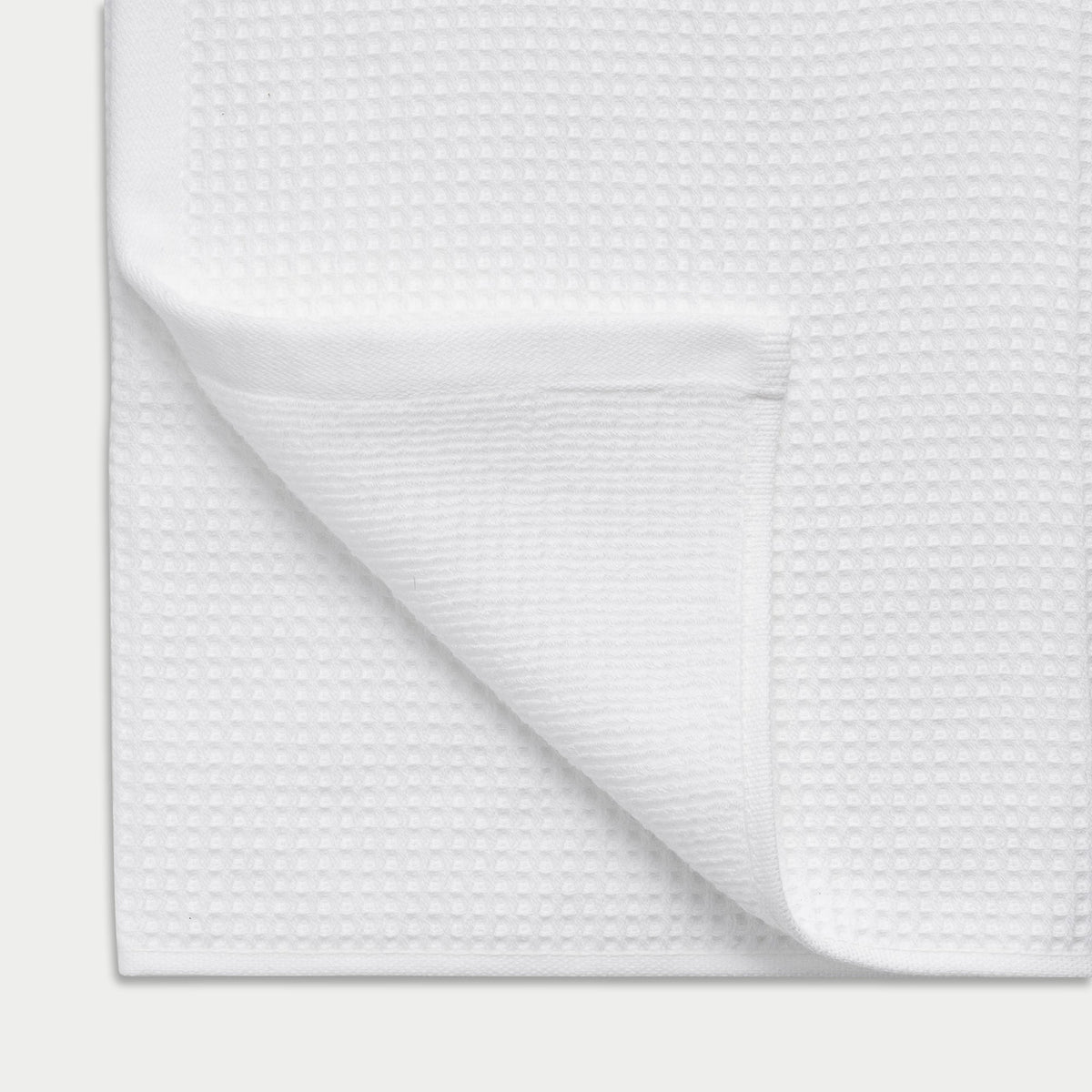 White Waffle Hand Towel resting flat on a white background. The corner is folded so that both sides of the towel can be seen. |Color:White