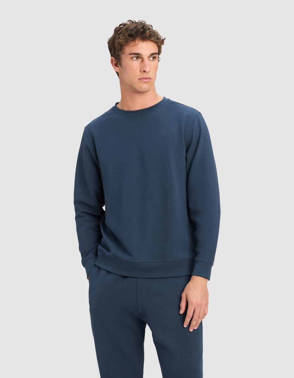 A young man wearing the Cozy Earth Men's StretchTech Crewneck in navy blue stands against a plain light gray background, looking to his left with one hand in his pocket.|Color:Eclipse