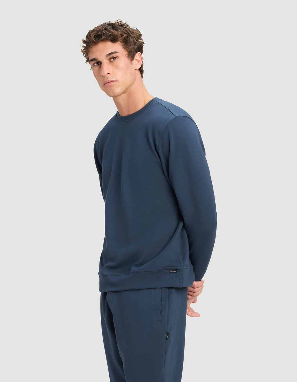 A young man with short, curly brown hair wears a Cozy Earth Men's StretchTech Crewneck in dark blue. He stands against a plain light grey background, looking slightly to the side with his hands behind his back.|Color:Eclipse