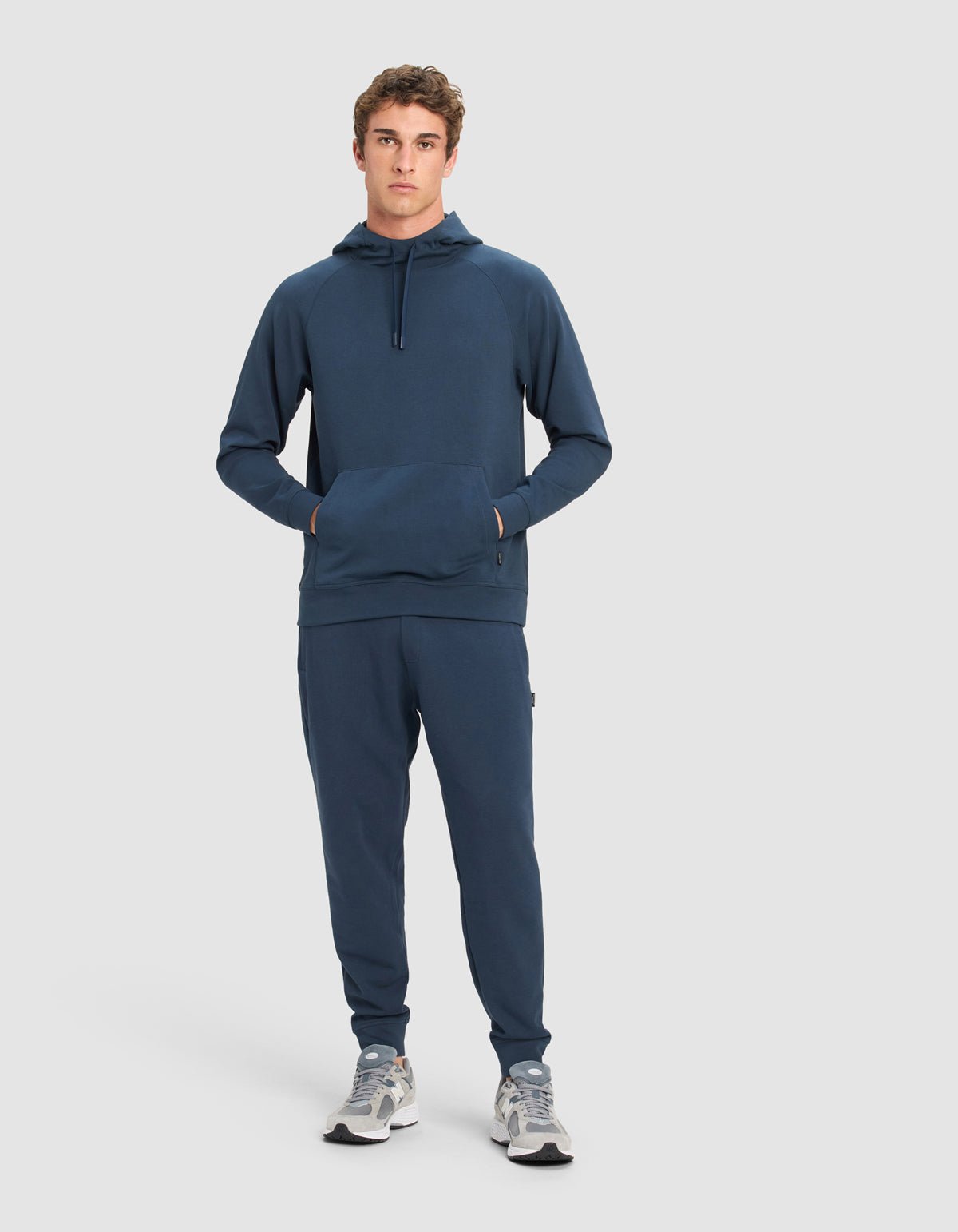 A young man stands facing forward, wearing a Cozy Earth Men's StretchTech Jogger set in dark blue with gray sneakers, hands in his hoodie pocket, against a plain light gray background.|Color:Eclipse
